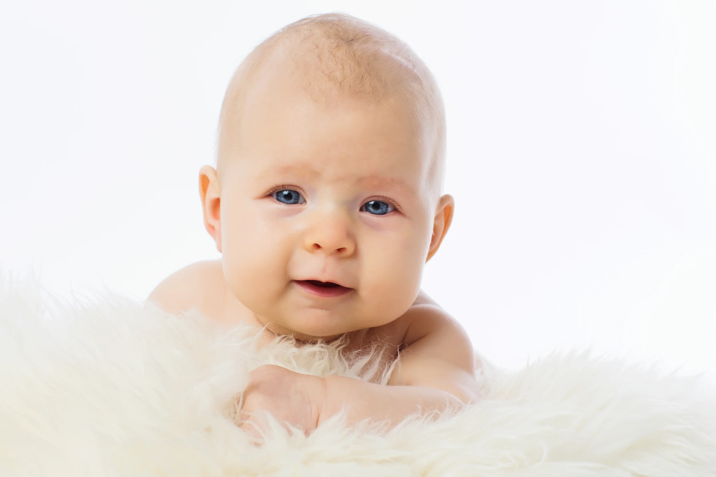 Blåøjet baby i babyfotografering, der ligger på hvidt lammefælde med mildt smil. Naturligt portræt.