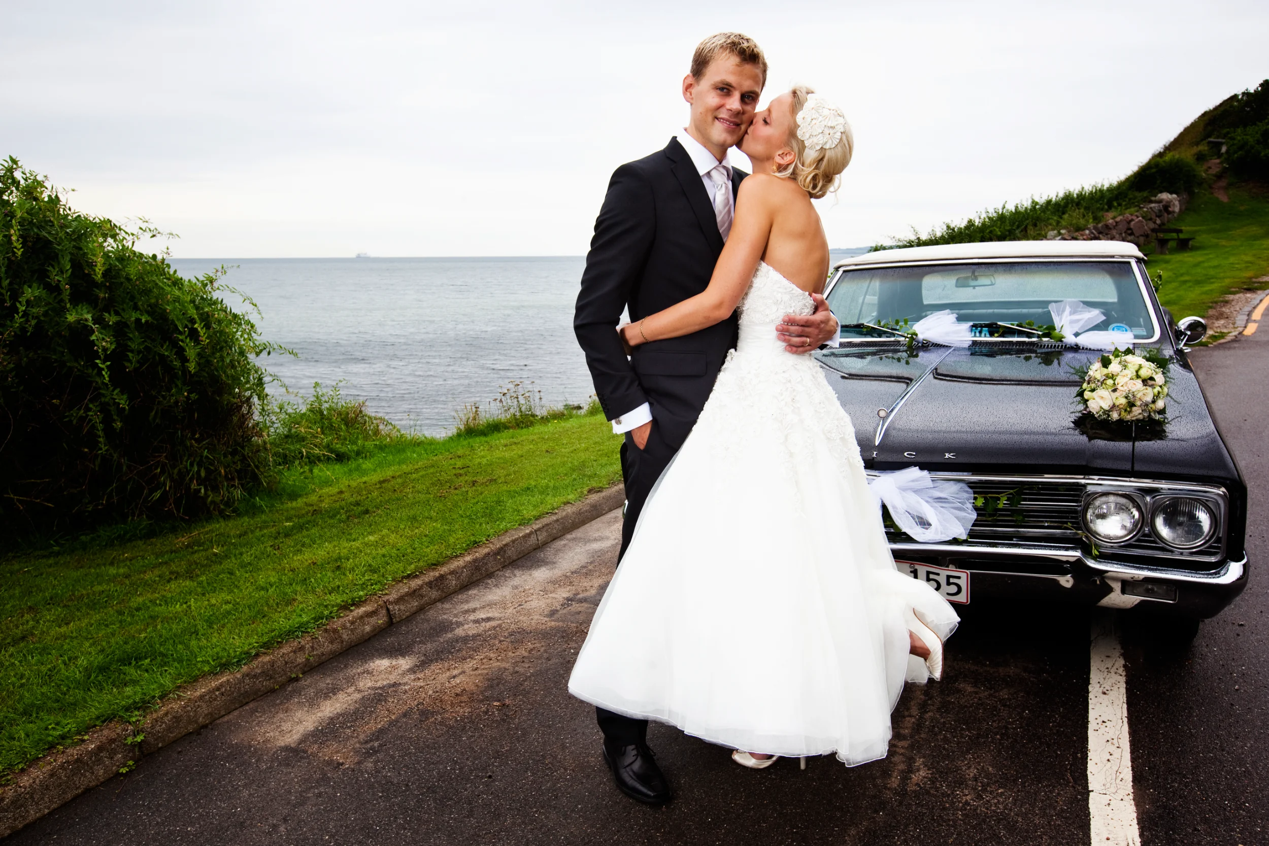 Bryllupsfotografering af brudepar, der kysser ved en sort veteranbil med havudsigt i baggrunden på en gråvejrsdag.