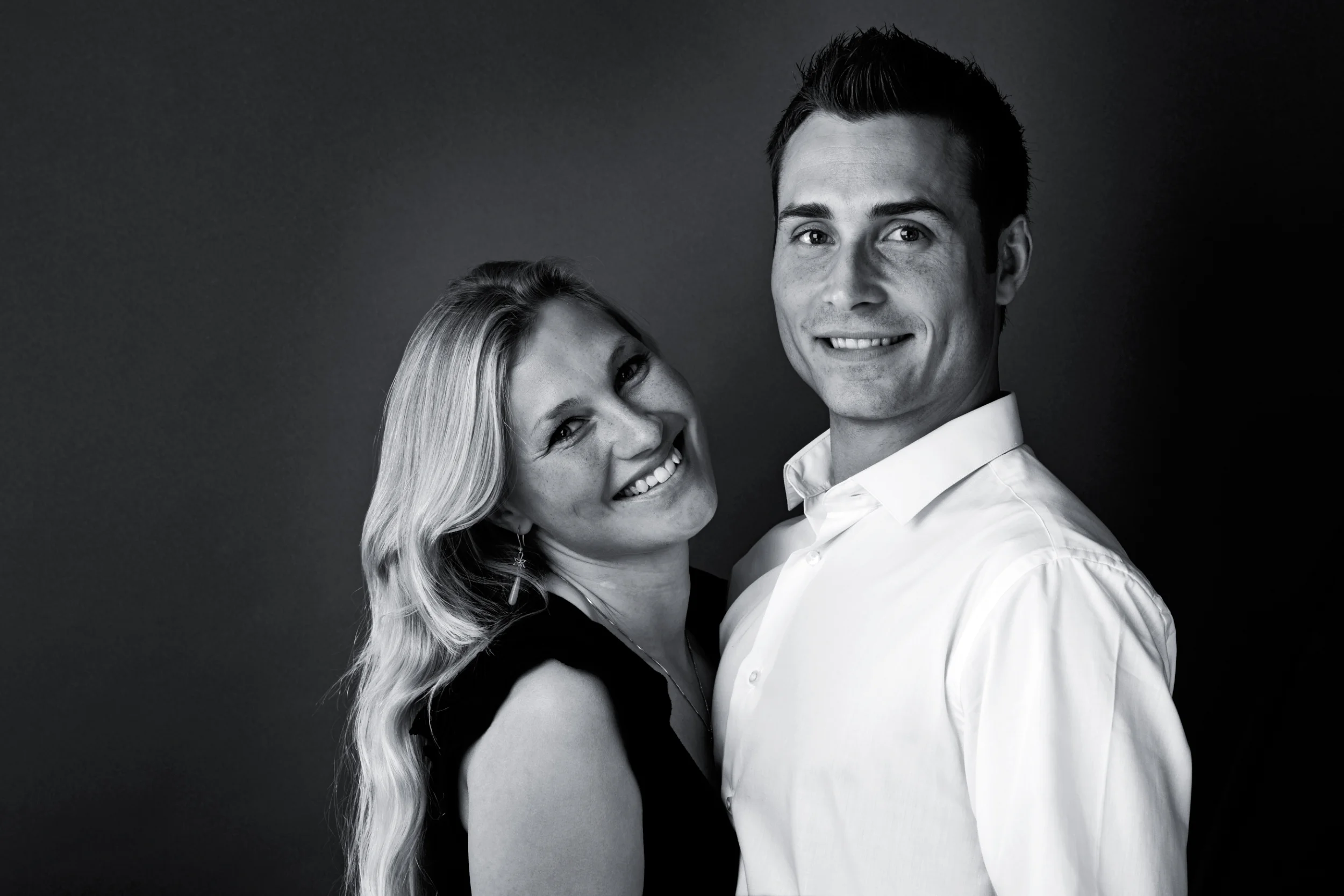 Professionelt sort-hvid portrætfoto af et smilende par, der står tæt med mørk baggrund og blødt lys.