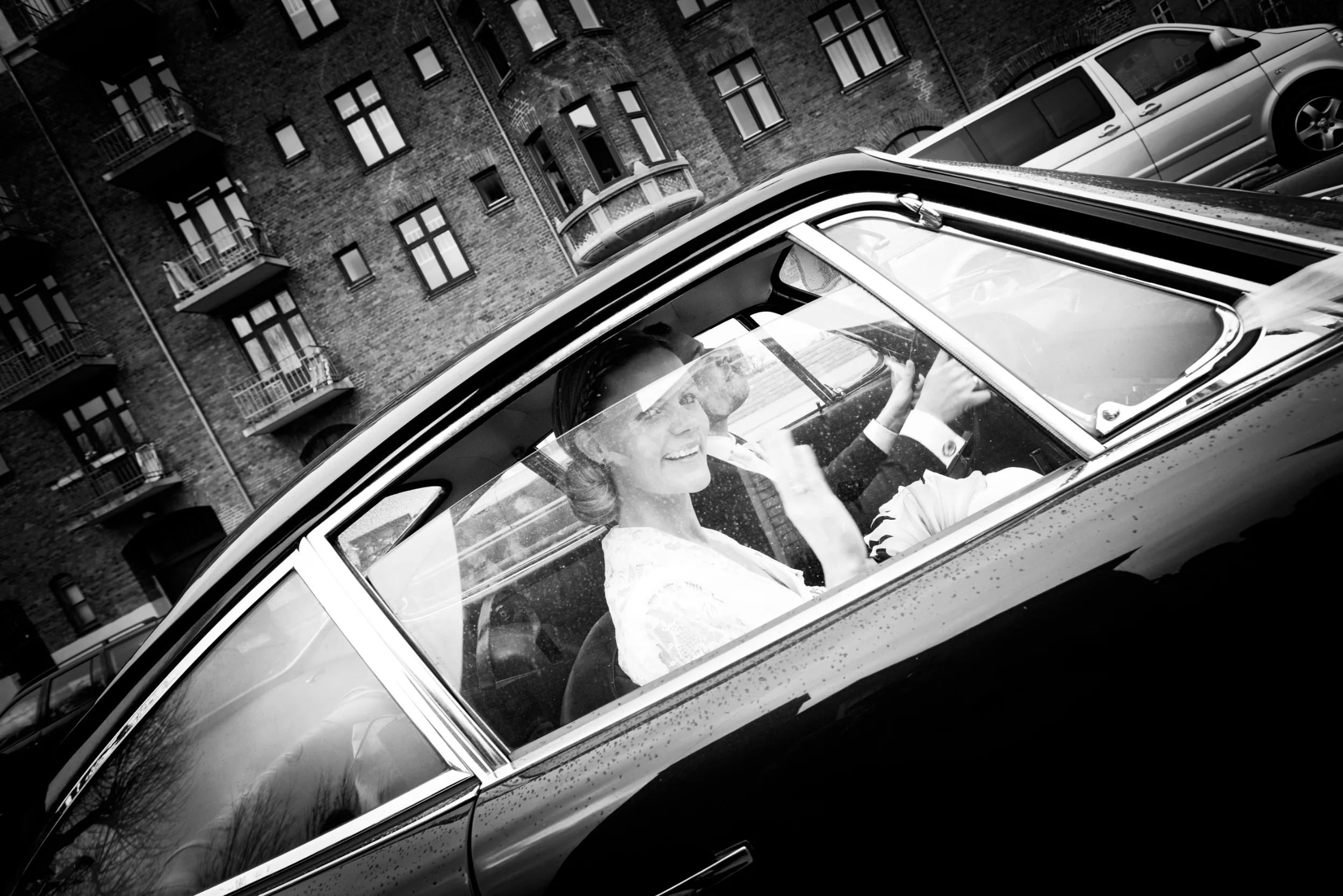 Bryllupsbillede af smilende brud med opsat hår, der vinker ud af vinduet på en sort bryllupsbil i sort/hvid