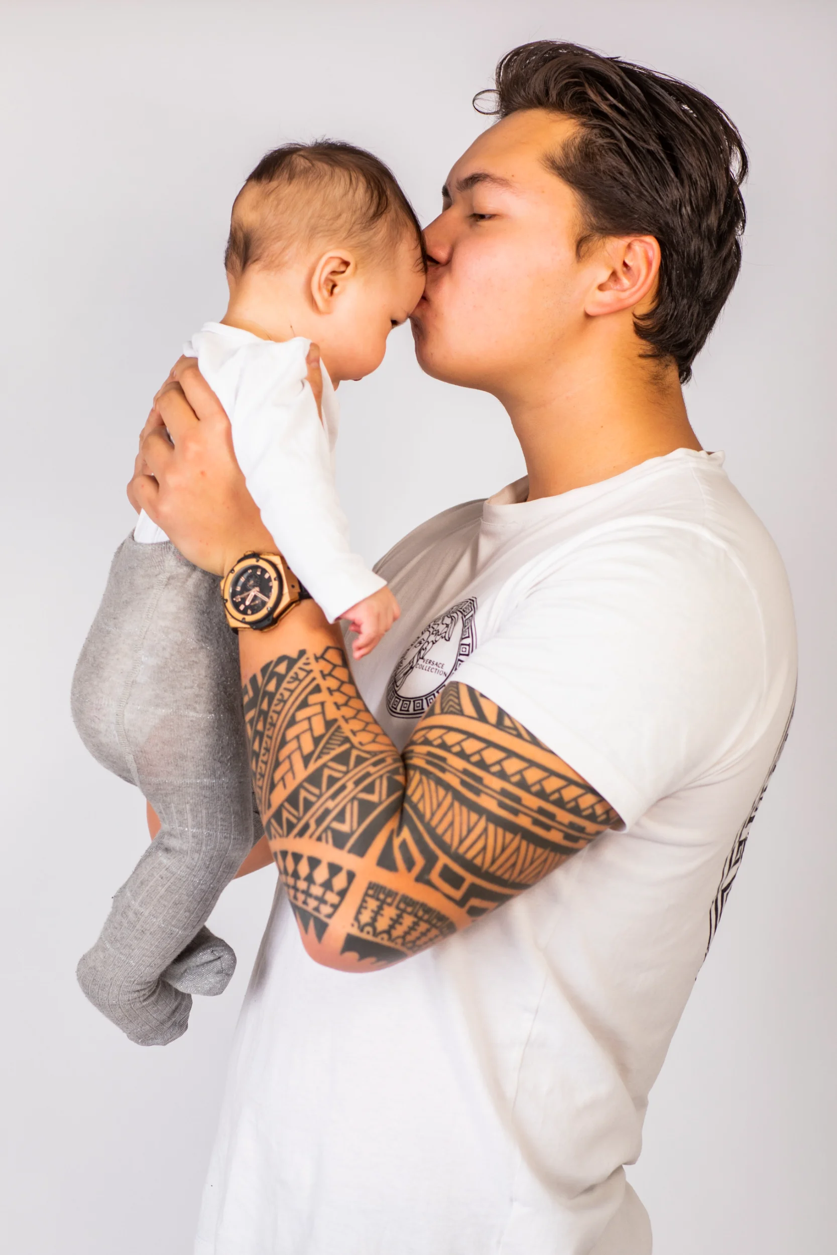 Nærbillede af far med tatoverede arme, der ømt kysser sin baby på panden. Et hjertevarmt babyfotografi i blødt lys.