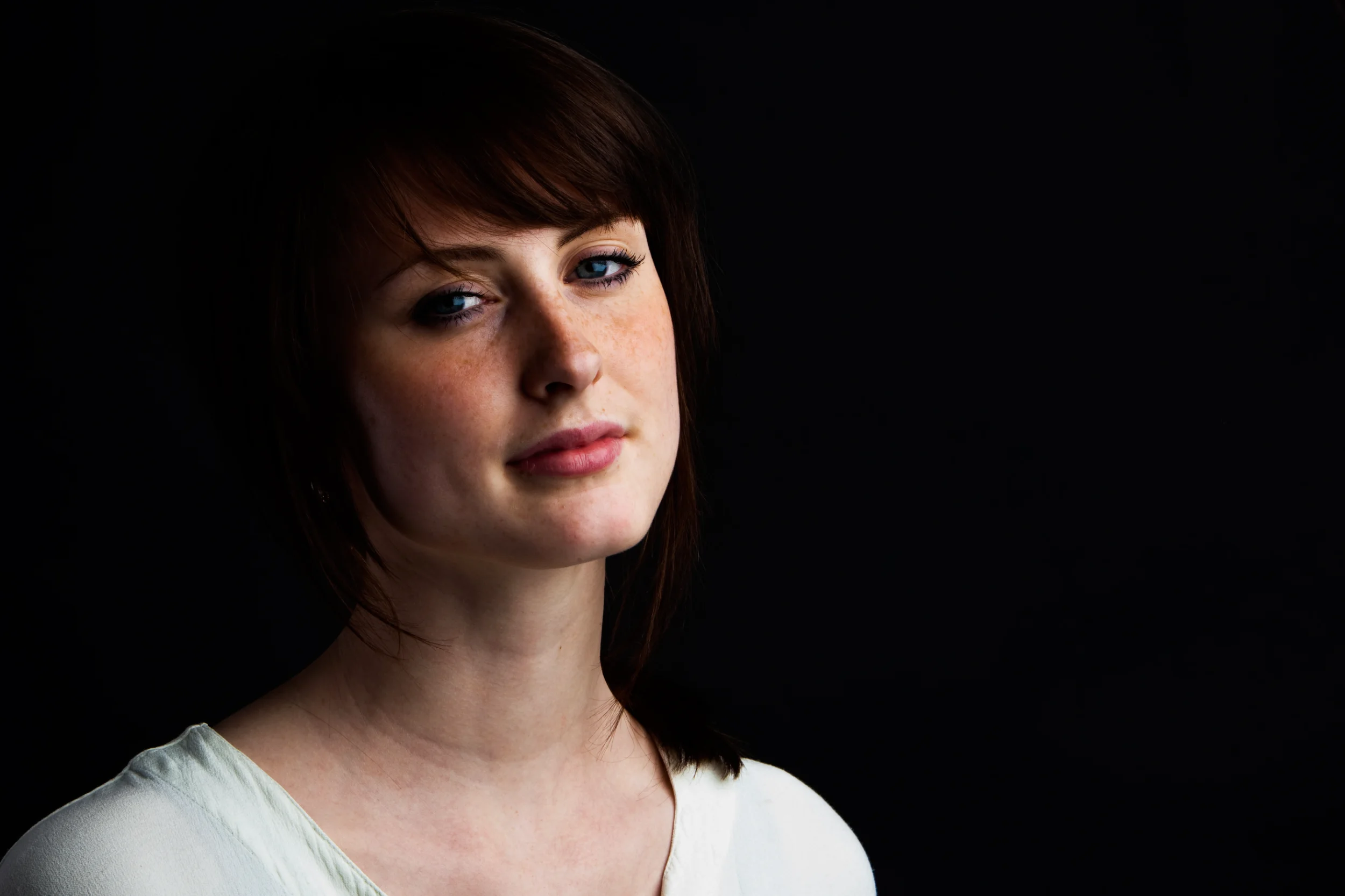 Portrætfoto af kvinde med fregner og brunt hår. Ansigtet er belyst af blødt, naturligt lys mod mørk baggrund.