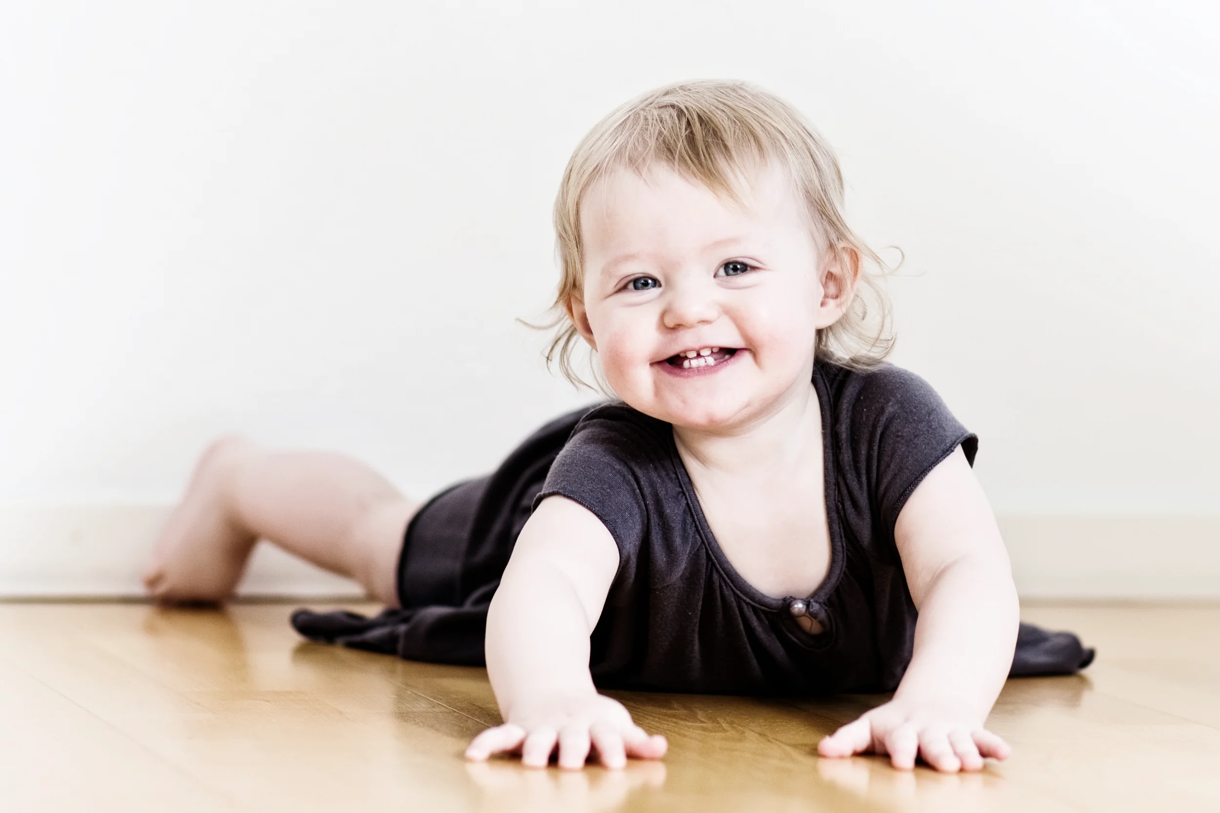 Glad baby kravler på trægulv med stort smil. Baby fotografering i lyse farver med naturligt lys.