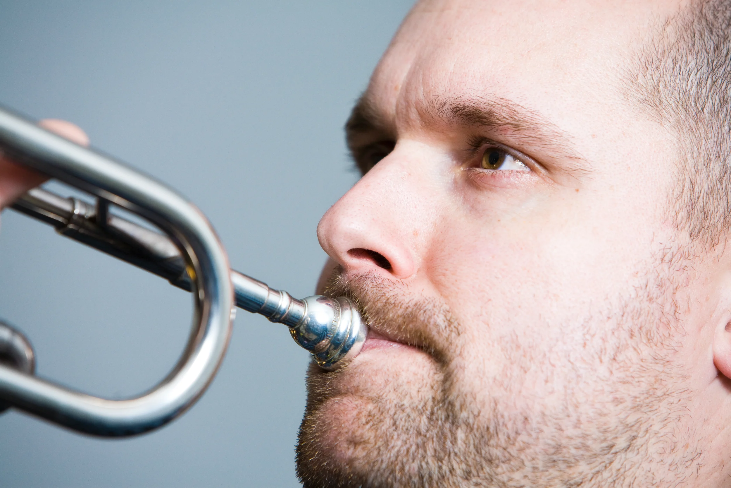 Nærbillede af mands ansigt der spiller på trompet; man ser mundstykket mod hans læber. Portrætfoto.