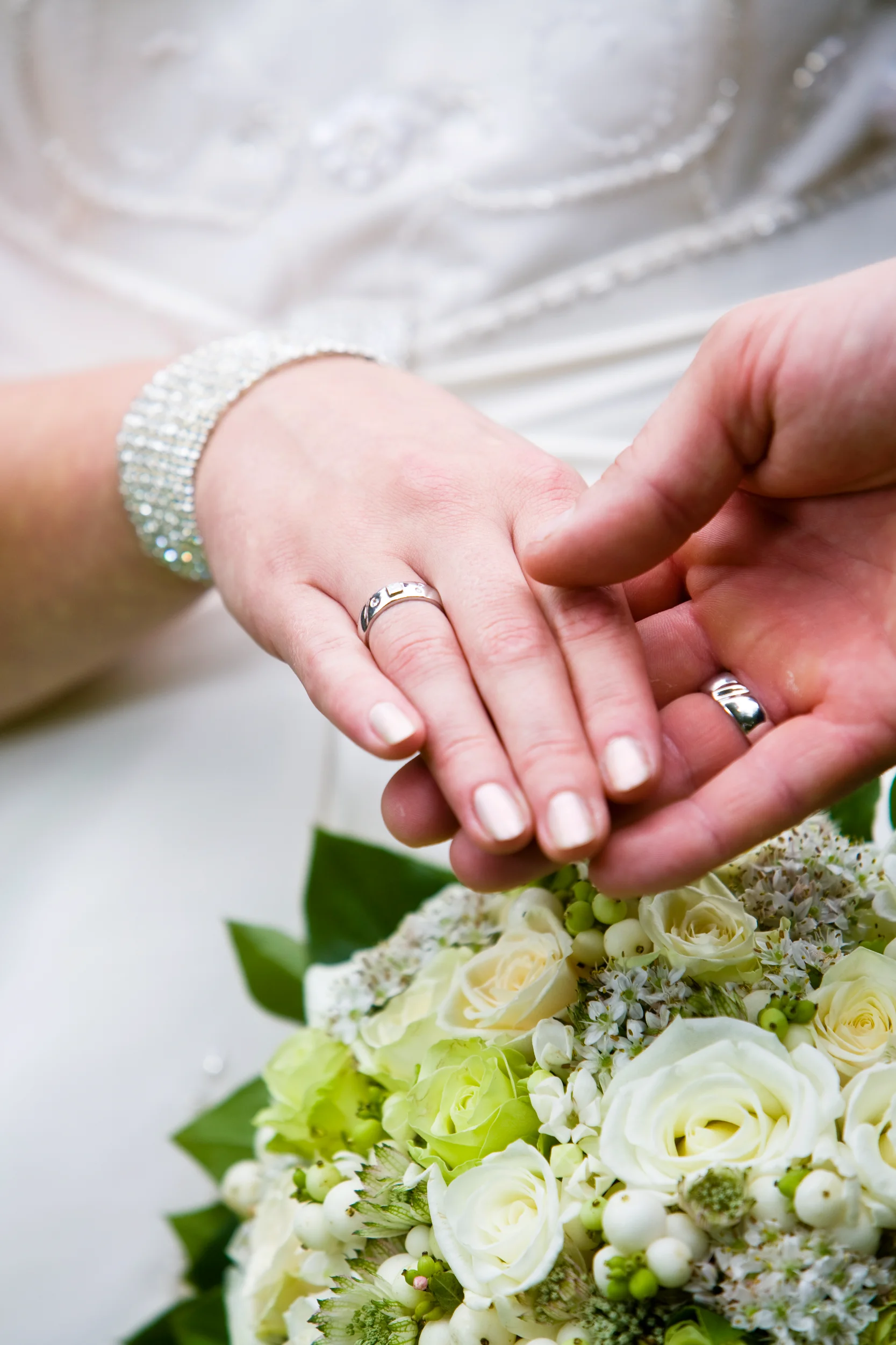 Bryllup: Nærbillede af brudeparrets hænder med vielsesringe, der rører ved en hvid og grøn brudebuket.