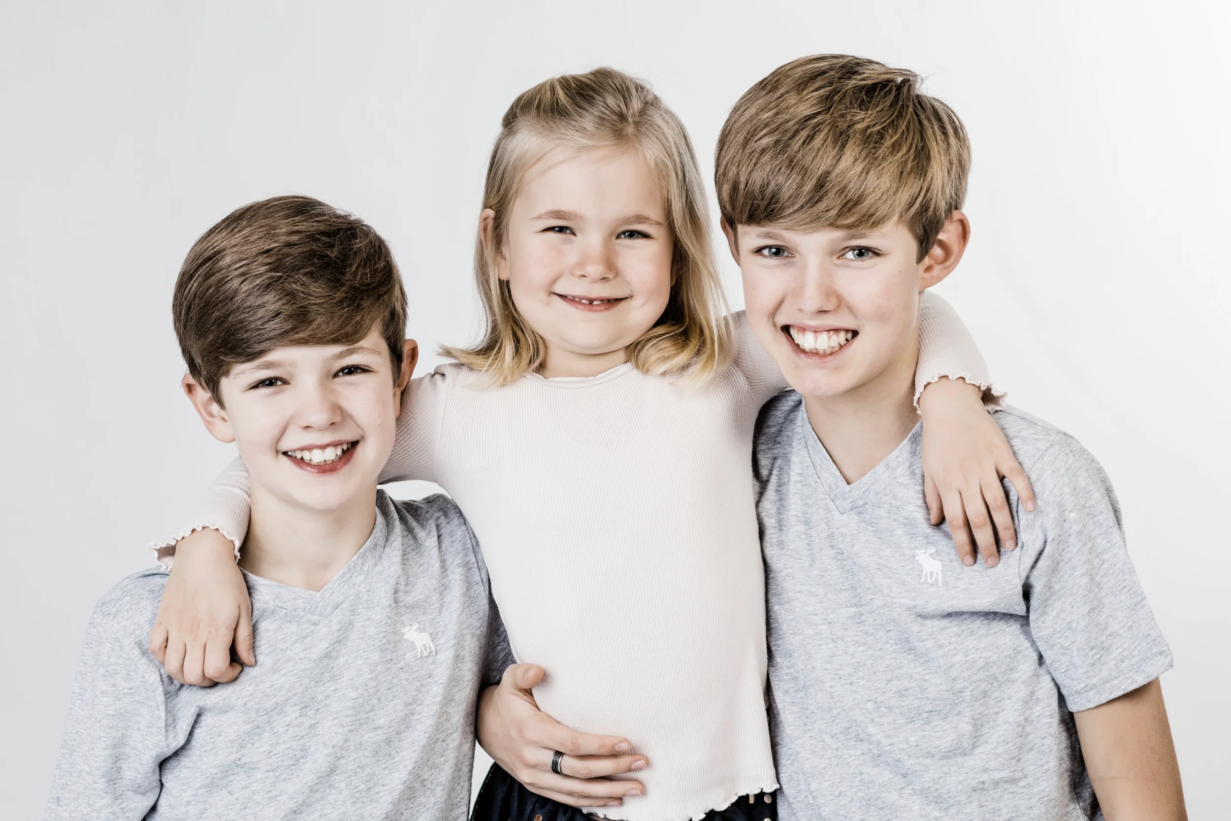 Tre glade børn i en børnefotografering, der smiler ind i kameraet med armene om hinanden på en hvid baggrund.