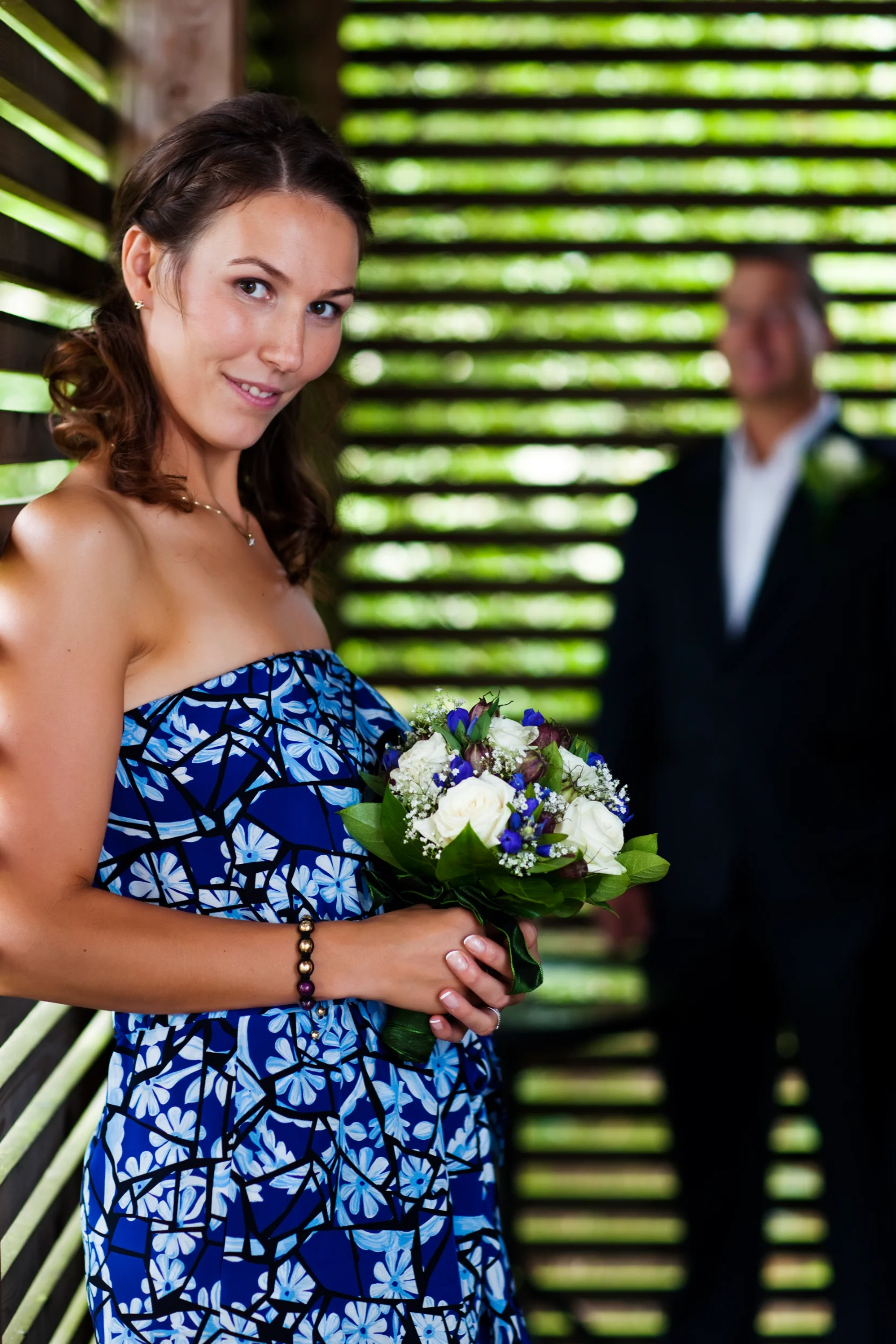 Glad bryllupsgæst i blå kjole med farverig blomsterbuket, en mand ses sløret bagved i naturligt lys.