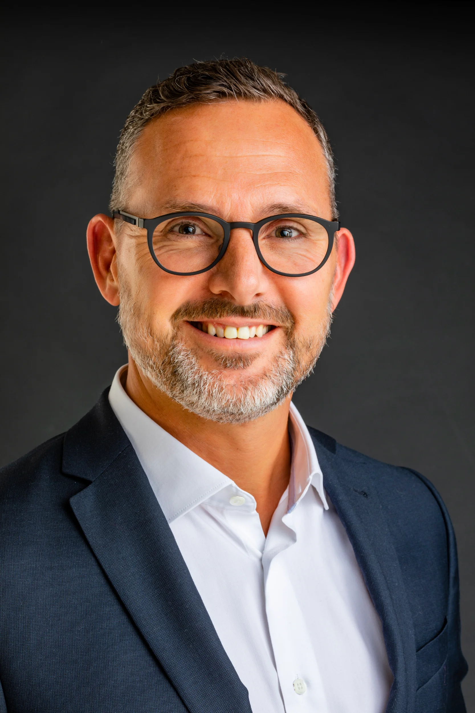 Portrætfoto af smilende mand med gråt skæg og briller, iført mørk jakke og hvid skjorte - varm belysning.