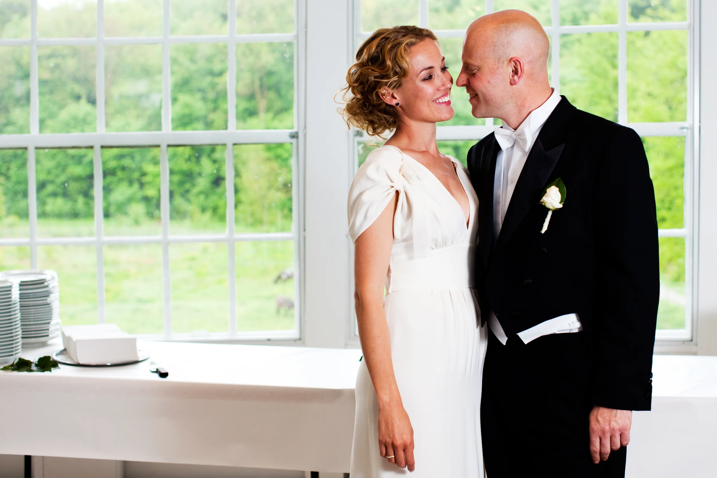 Bryllupsfotografering: Smilende brudepar i naturligt lys foran et stort vindue med grønne træer udenfor.