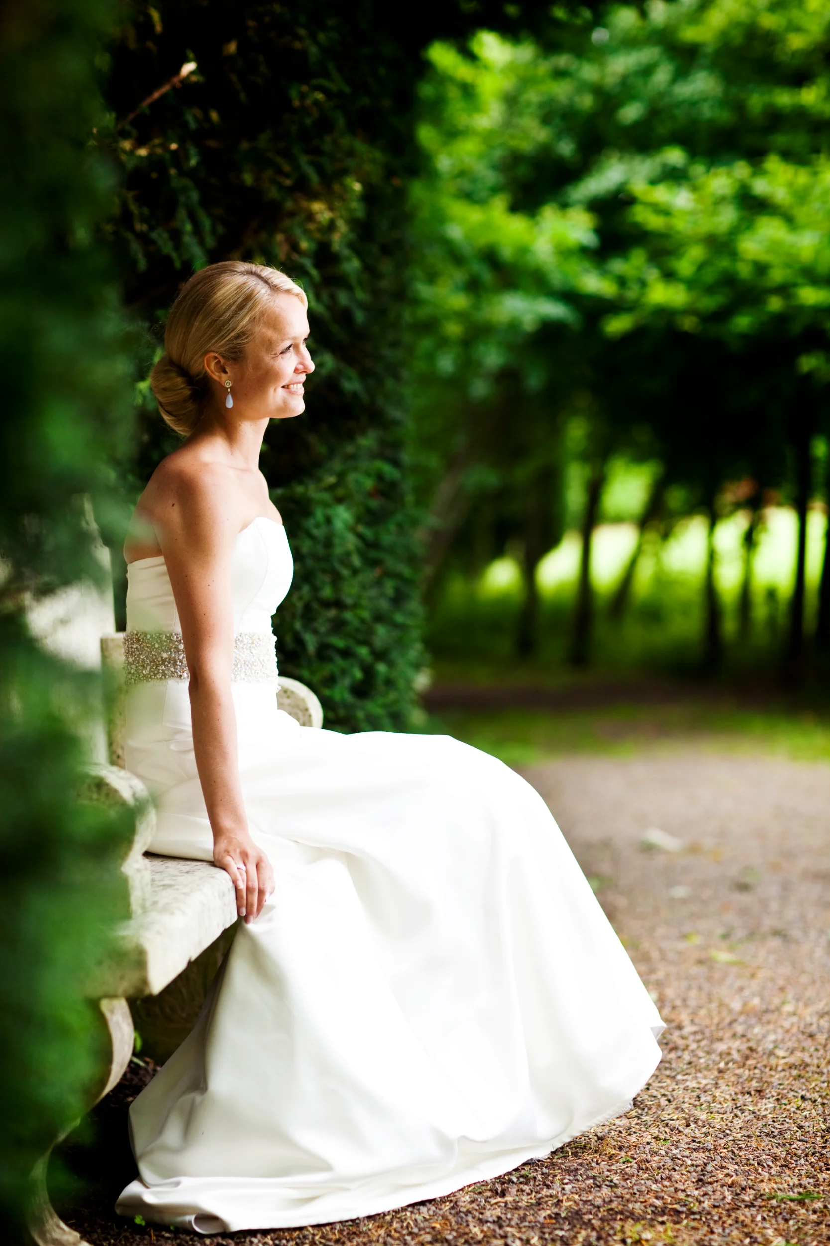 Brud i hvid kjole sidder smilende på stenbænk i grøn have. Et autentisk bryllupsfoto i naturligt lys.