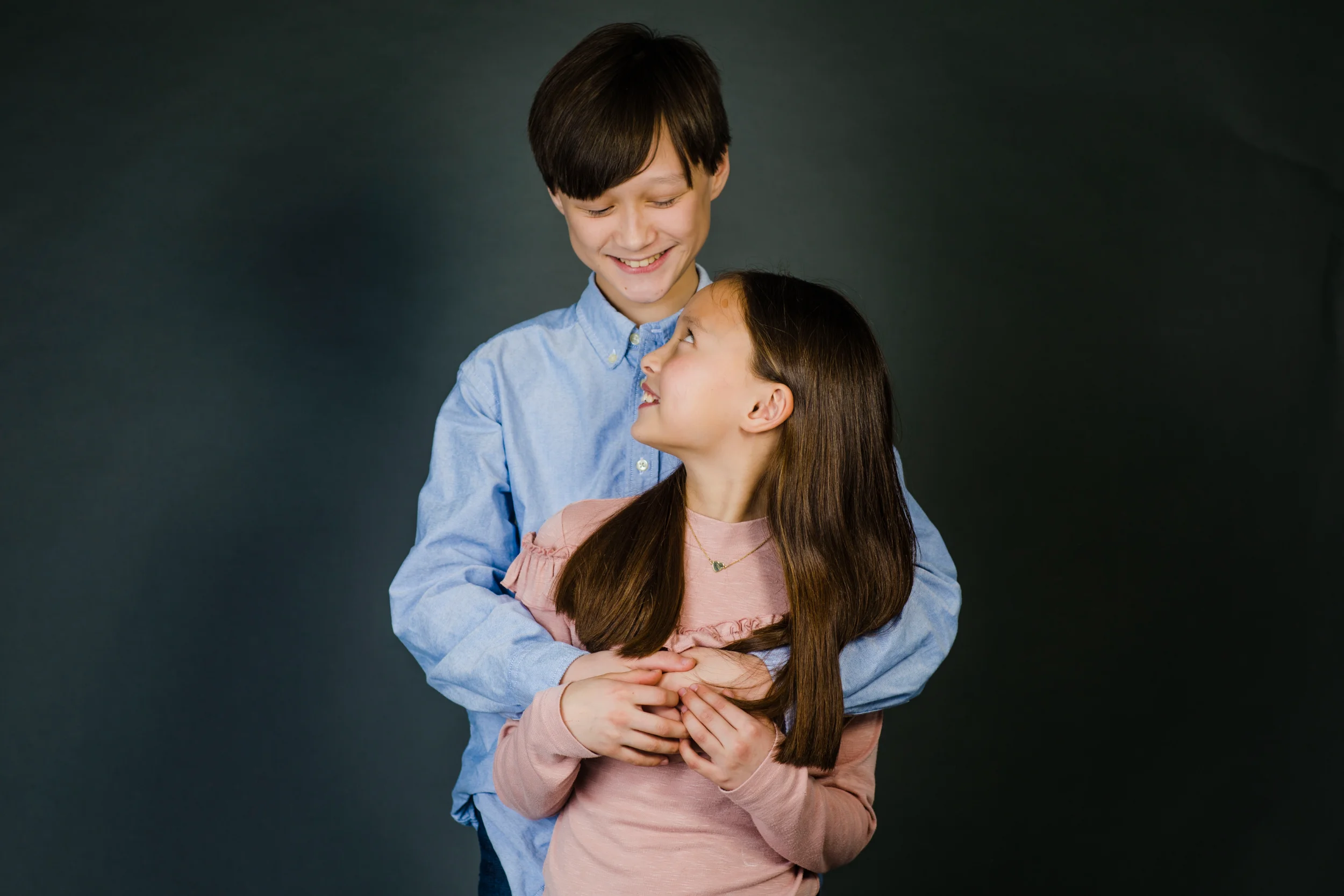 Søskendefoto med en dreng, der omfavner sin lillesøster bagfra. De smiler, og hun kigger op på ham med glæde.