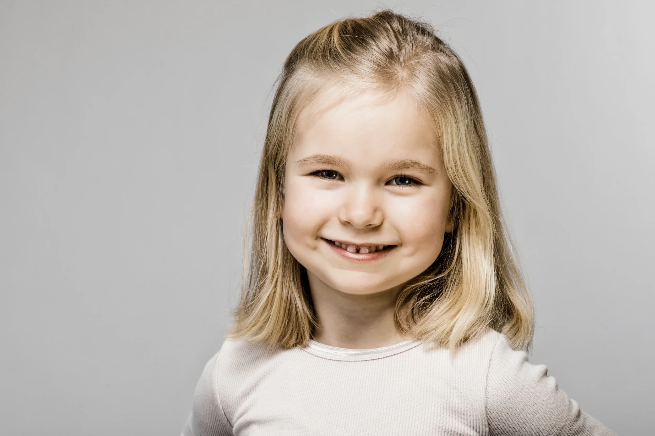 Nært portrætfoto af en glad pige med blondt hår, der smiler naturligt mod beskueren. Et charmerende børnefoto.