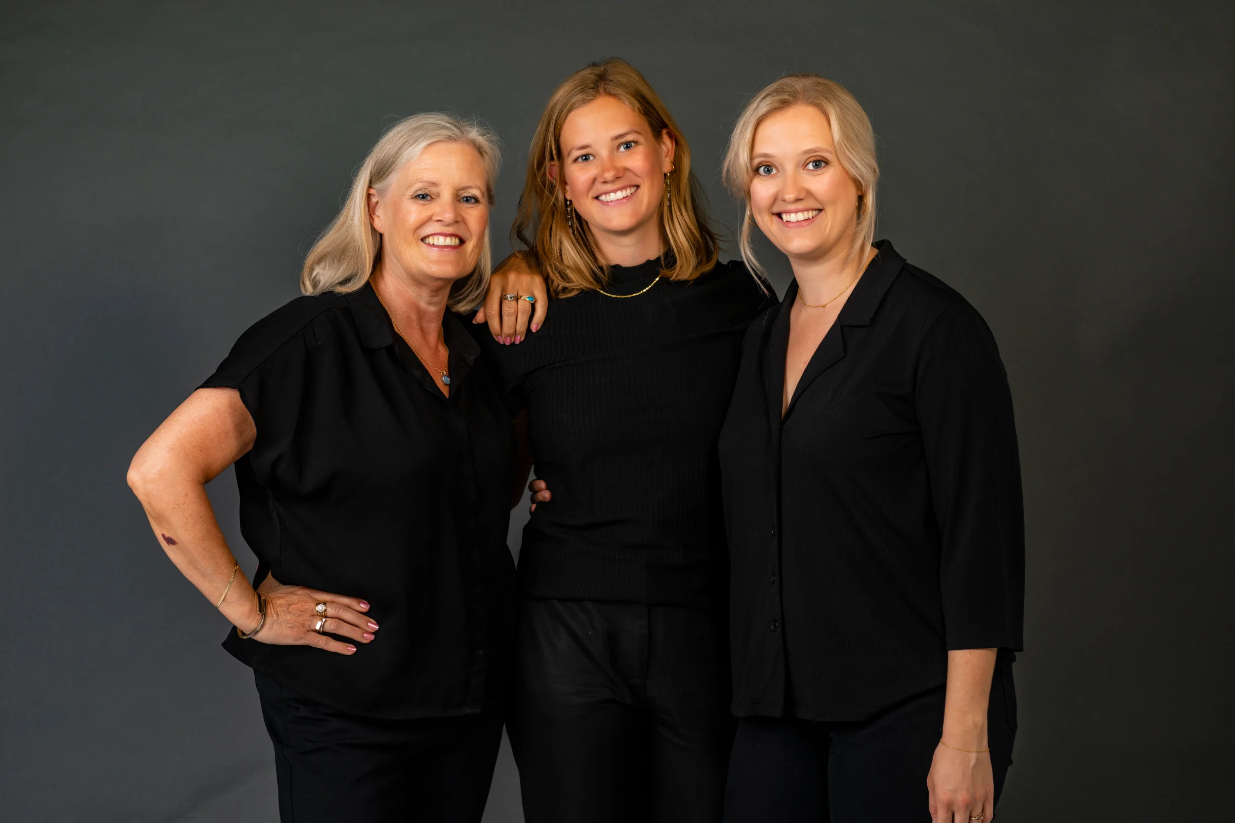 Glad familie fotograferet i sort tøj mod mørk baggrund. Midaldrende kvinde med to yngre, smilende kvinder.