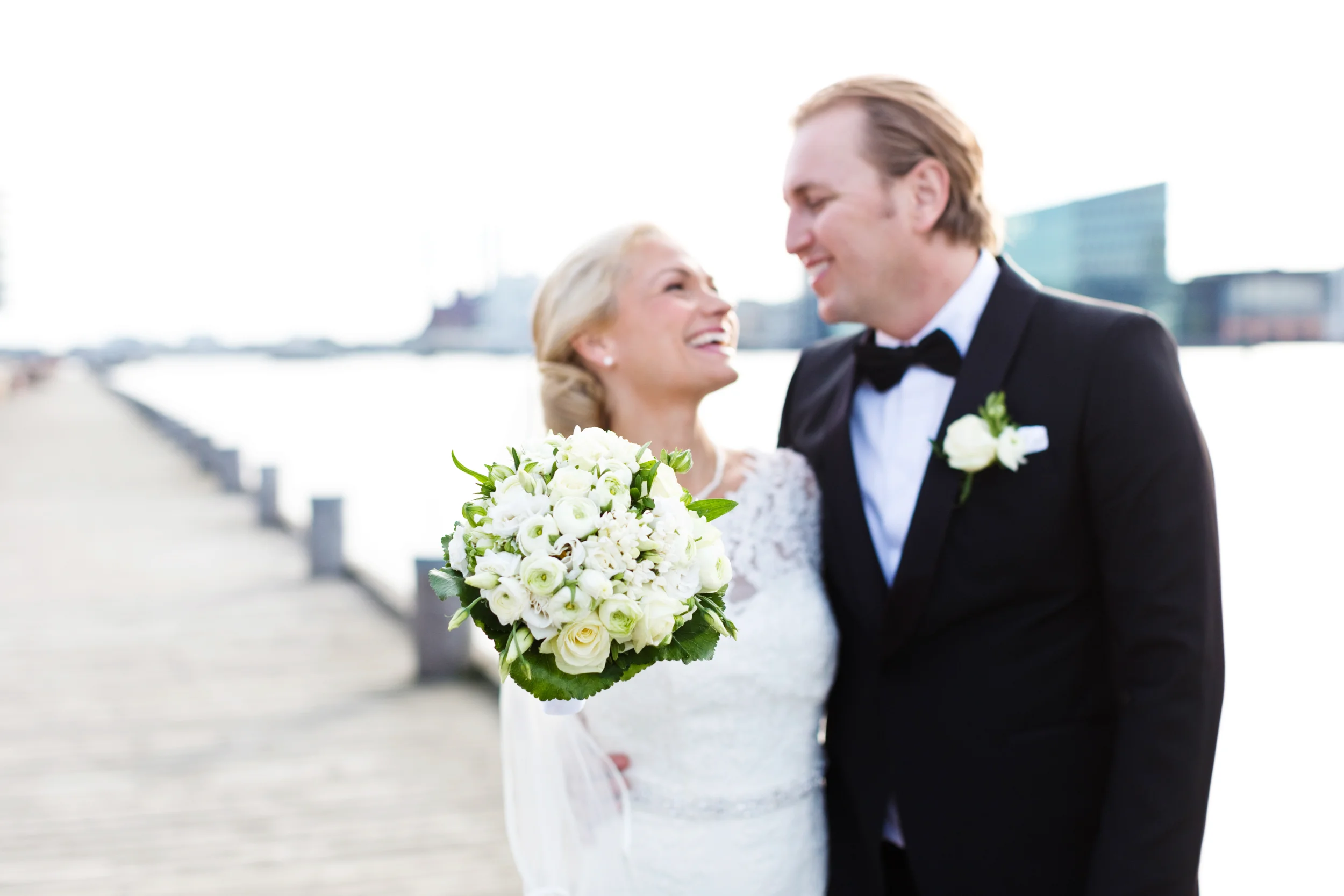 Bryllupsfoto af smilende brudepar ved vandet, med bruden holdende en hvid buket i blødt dagslys.