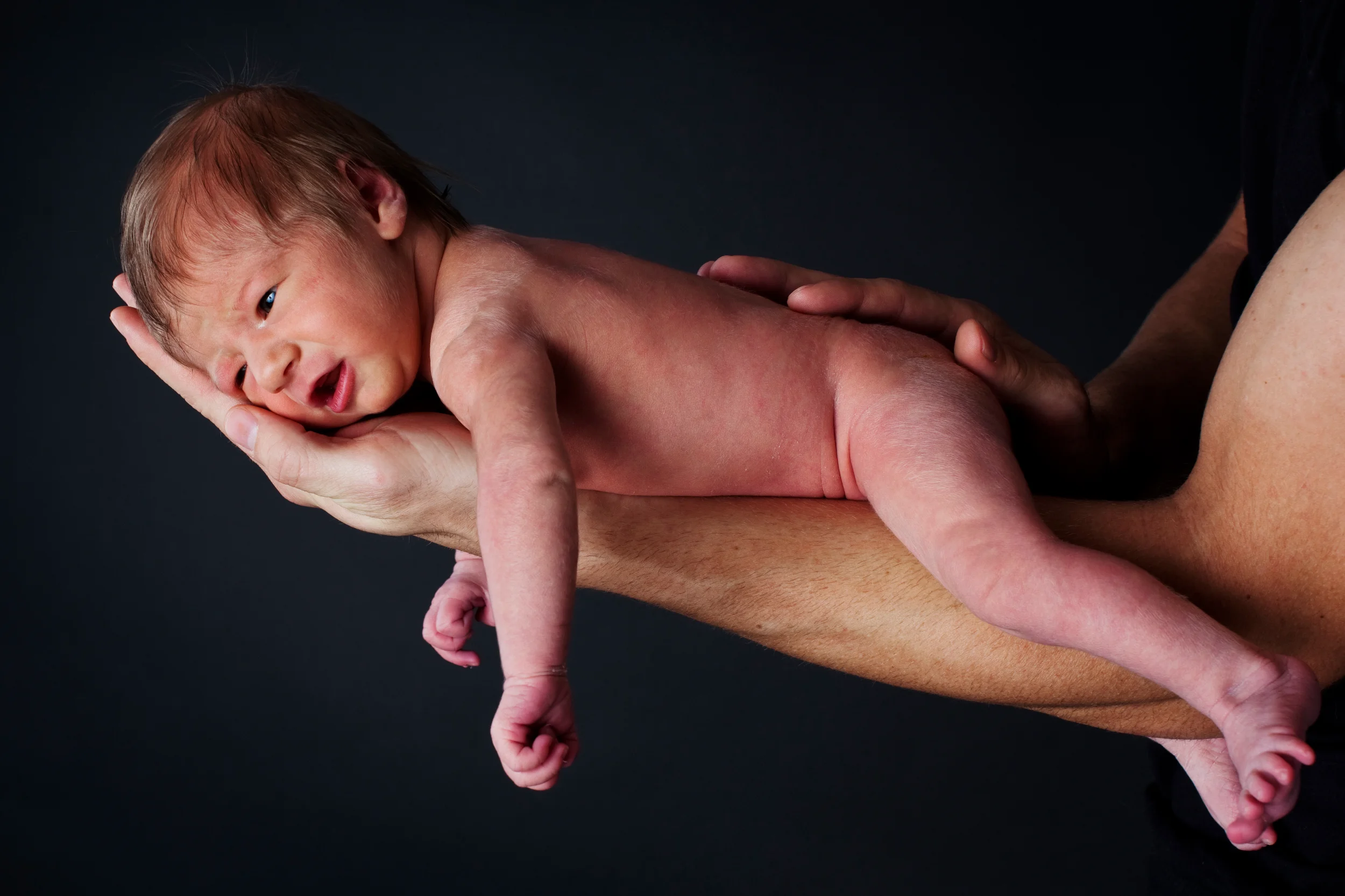Babyfotografering i sort/hvid af en nyfødt baby liggende på en voksens hænder med mørk baggrund.