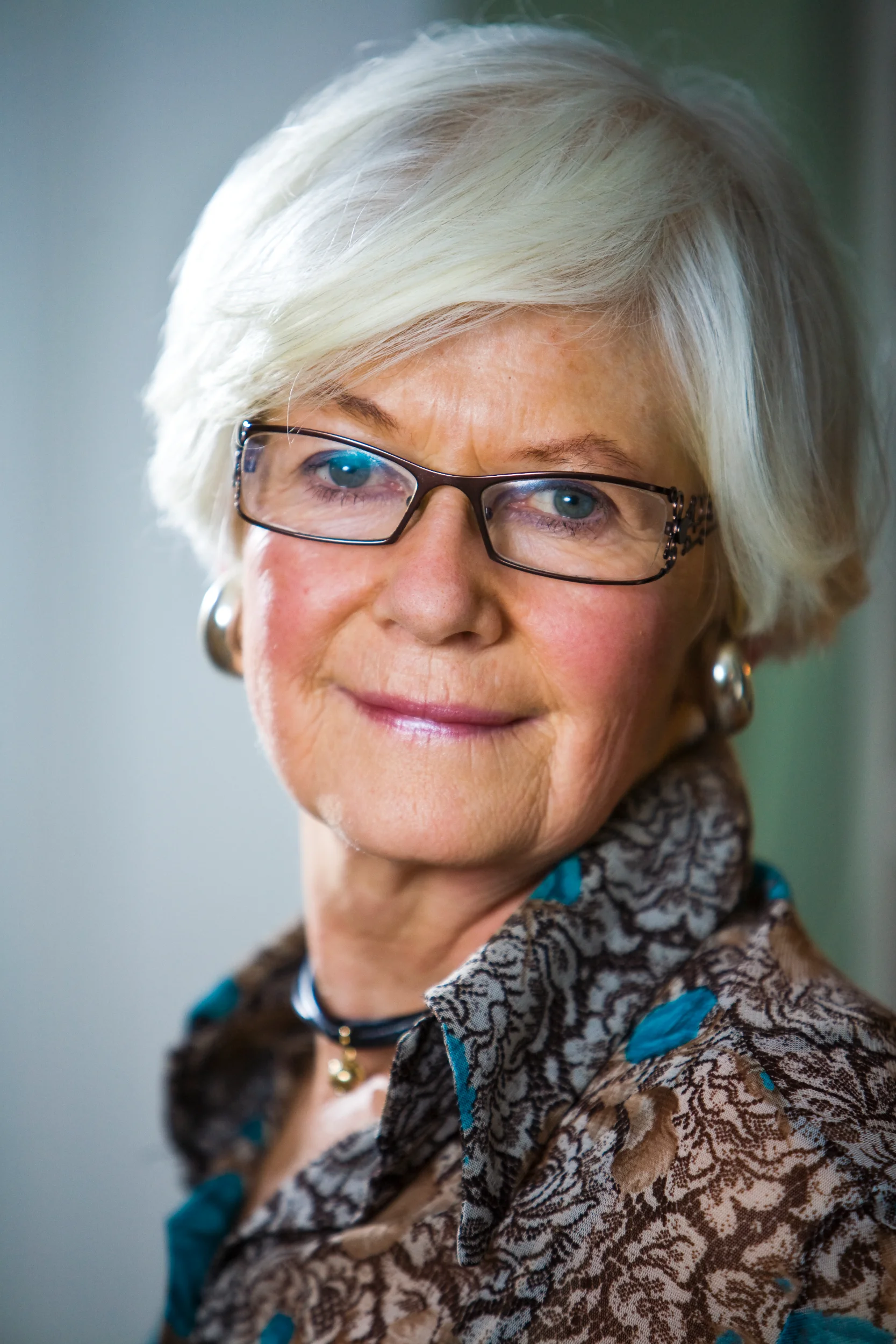 Portrætfoto af smilende ældre kvinde med kort, hvidt hår og briller. Blidt lys fremhæver hendes venlige udtryk.