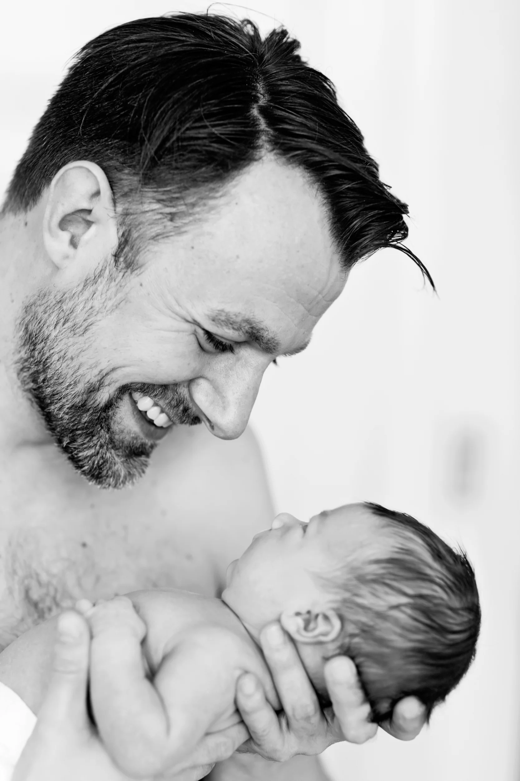 Babyfotografering i sort/hvid af nybagt far, der smiler kærligt til sin baby i blødt sidelys. Intim stemning.