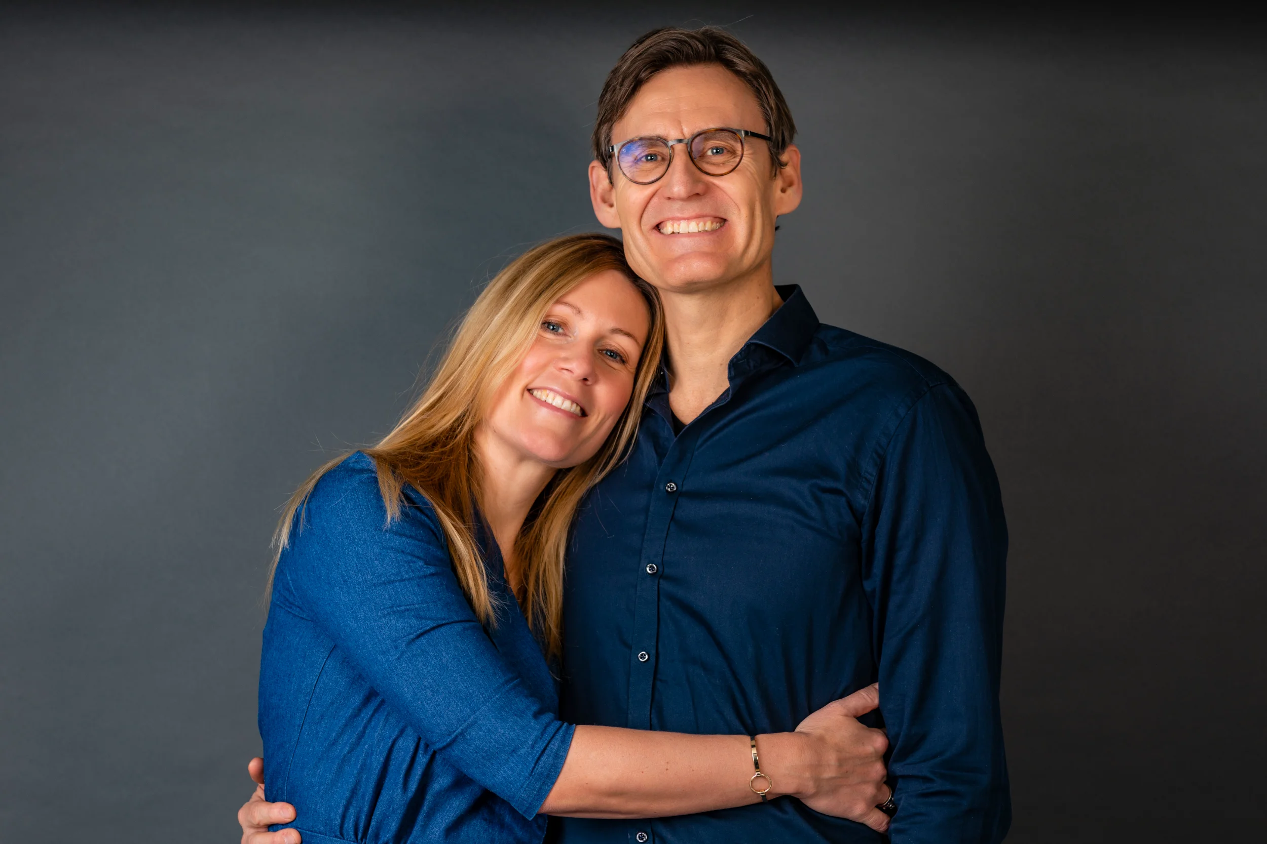 Glad kvinde med langt, lyst hår læner hovedet mod smilende mand i briller. Varmt portrætfoto med grå baggrund.