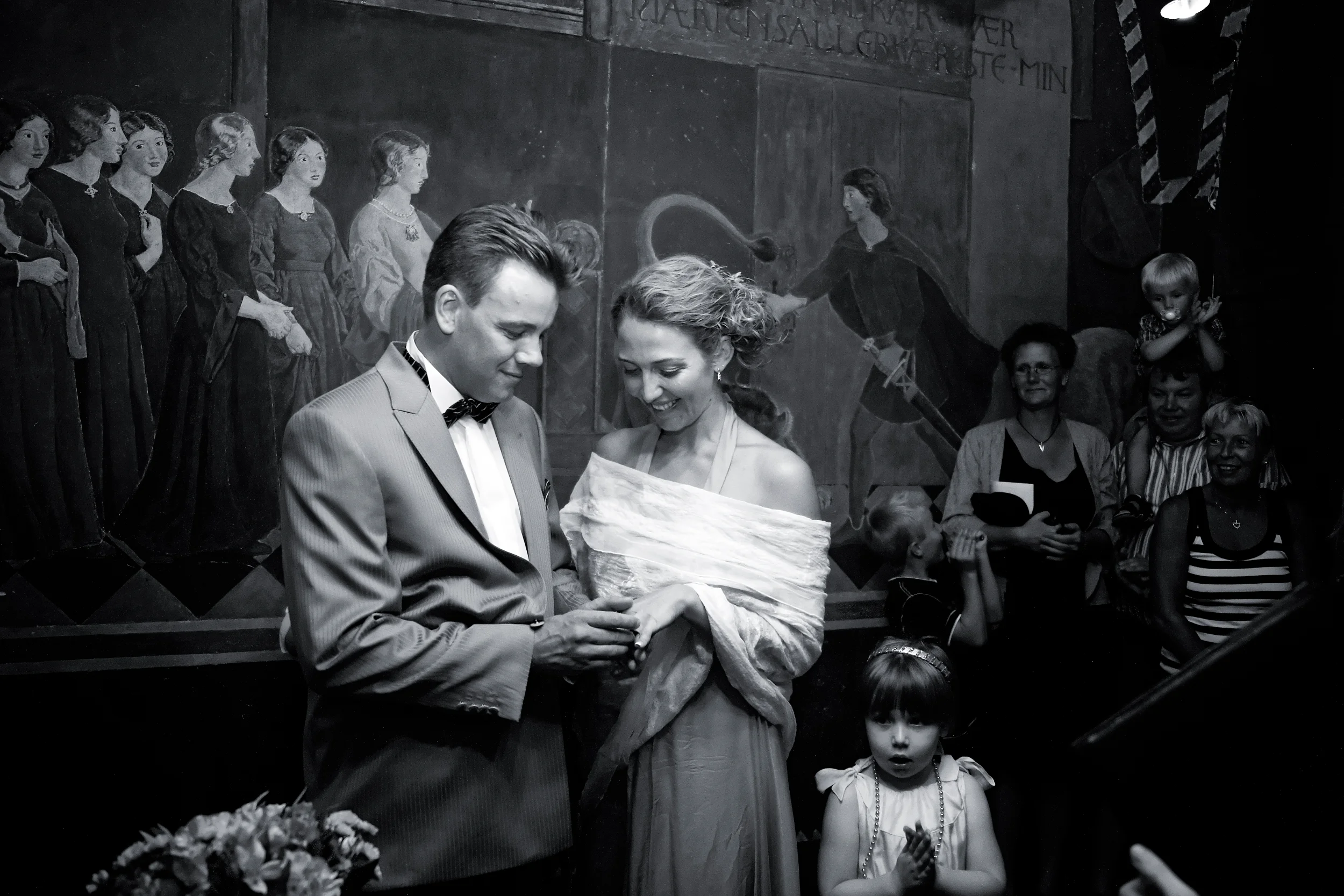 Bryllupsfotografering: Brudgom sætter ring på brudens finger i et rådhus med historiske vægmalerier i baggrunden.