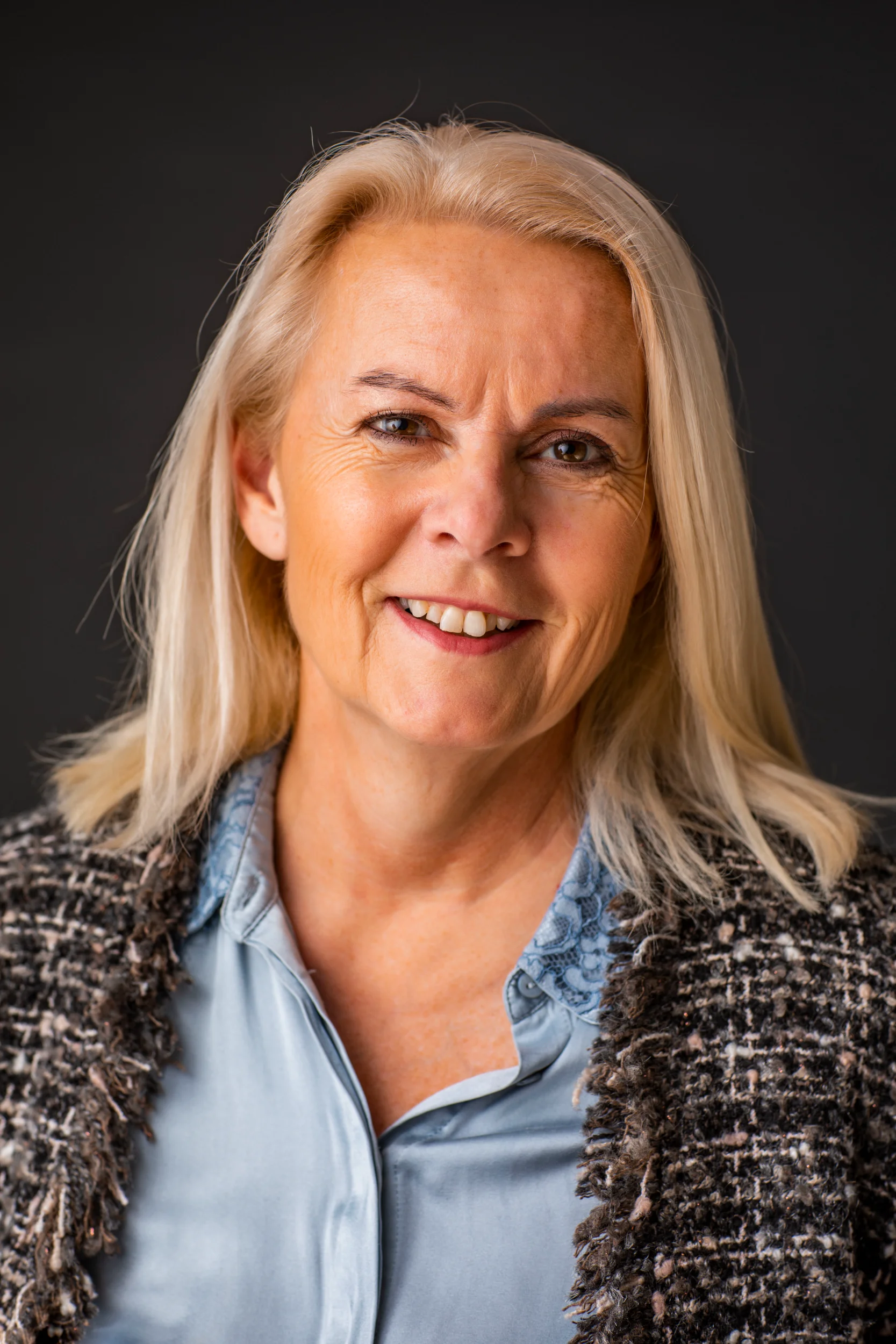 Portrætfoto af smilende kvinde med blondt hår i Virum, iført tweedjakke og blå skjorte mod mørk baggrund.
