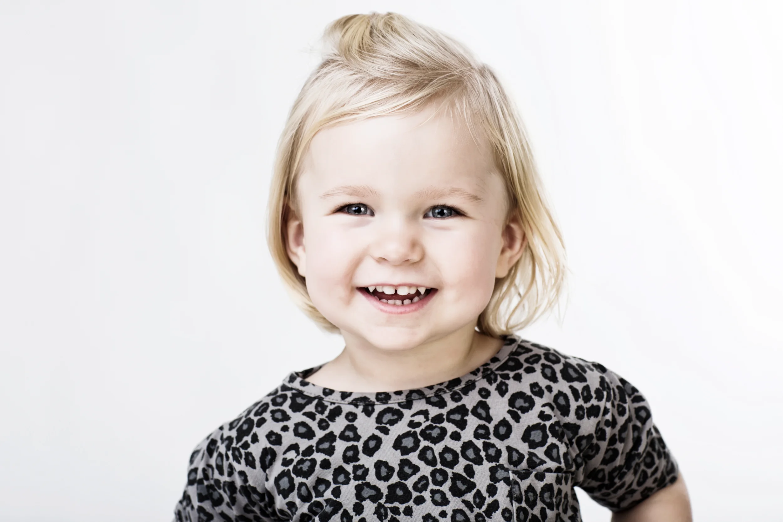 Børnefotografi af en smilende lille pige med lyst hår og leopardprint bluse mod hvid baggrund i blødt lys.