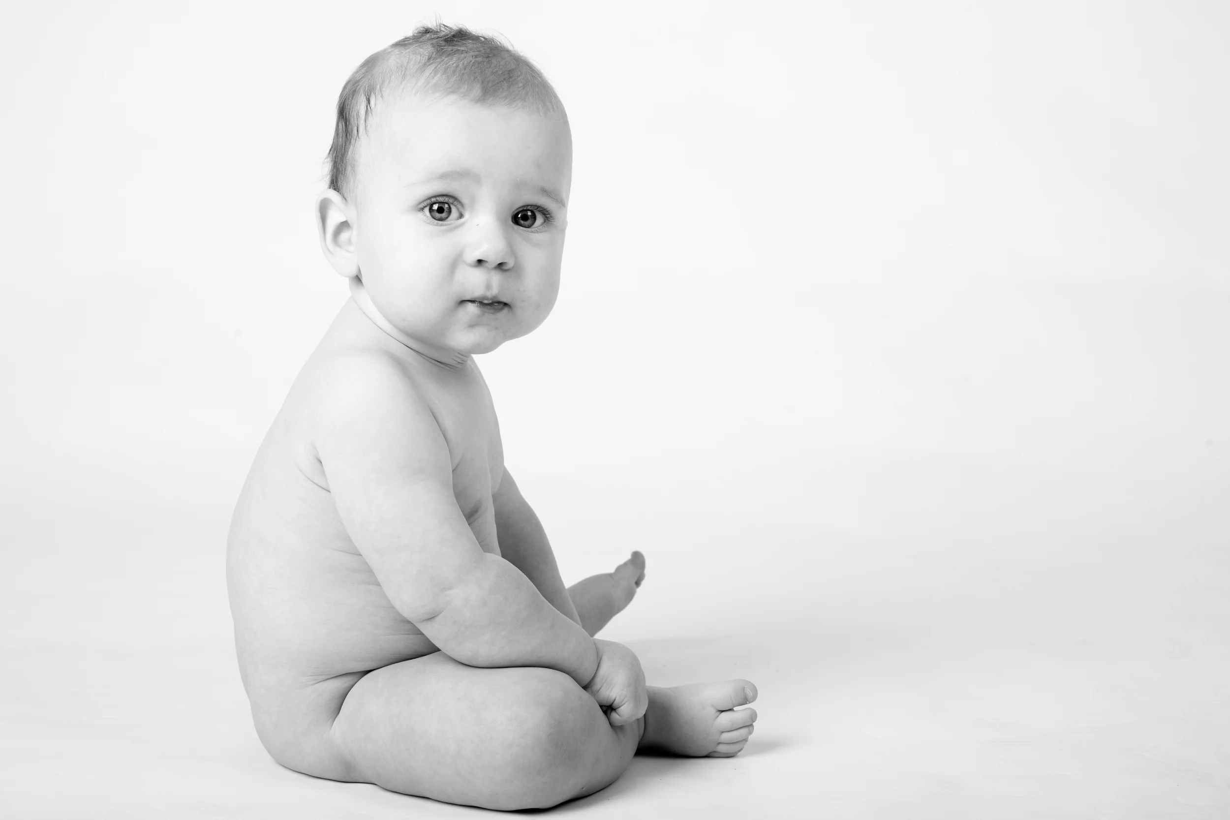 Sødt babyfoto af nøgen siddende baby med blå øjne, der kigger nysgerrigt mod kameraet på en hvid baggrund.