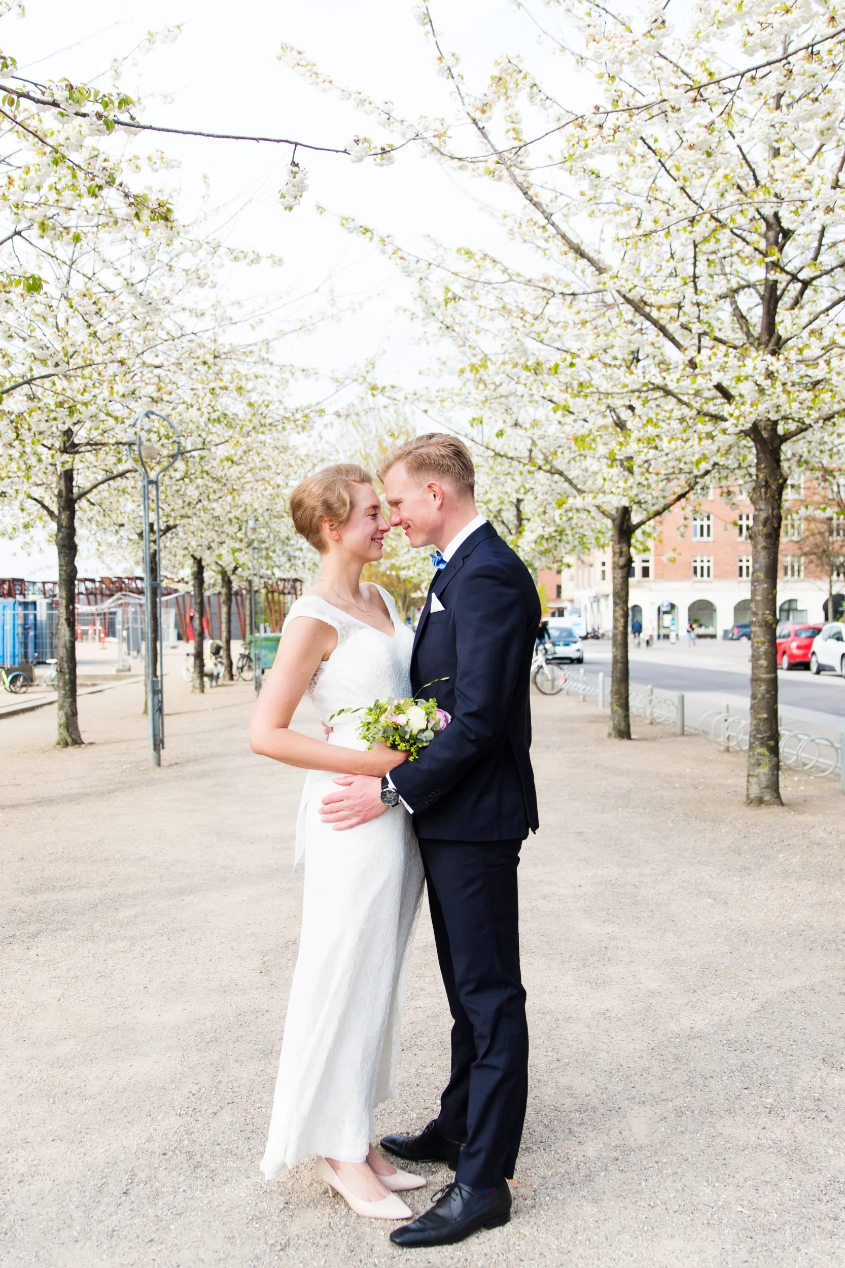 Bryllupsfoto af nygift par tæt omfavnende under florlette, hvide kirsebærtræer på en lys dag.