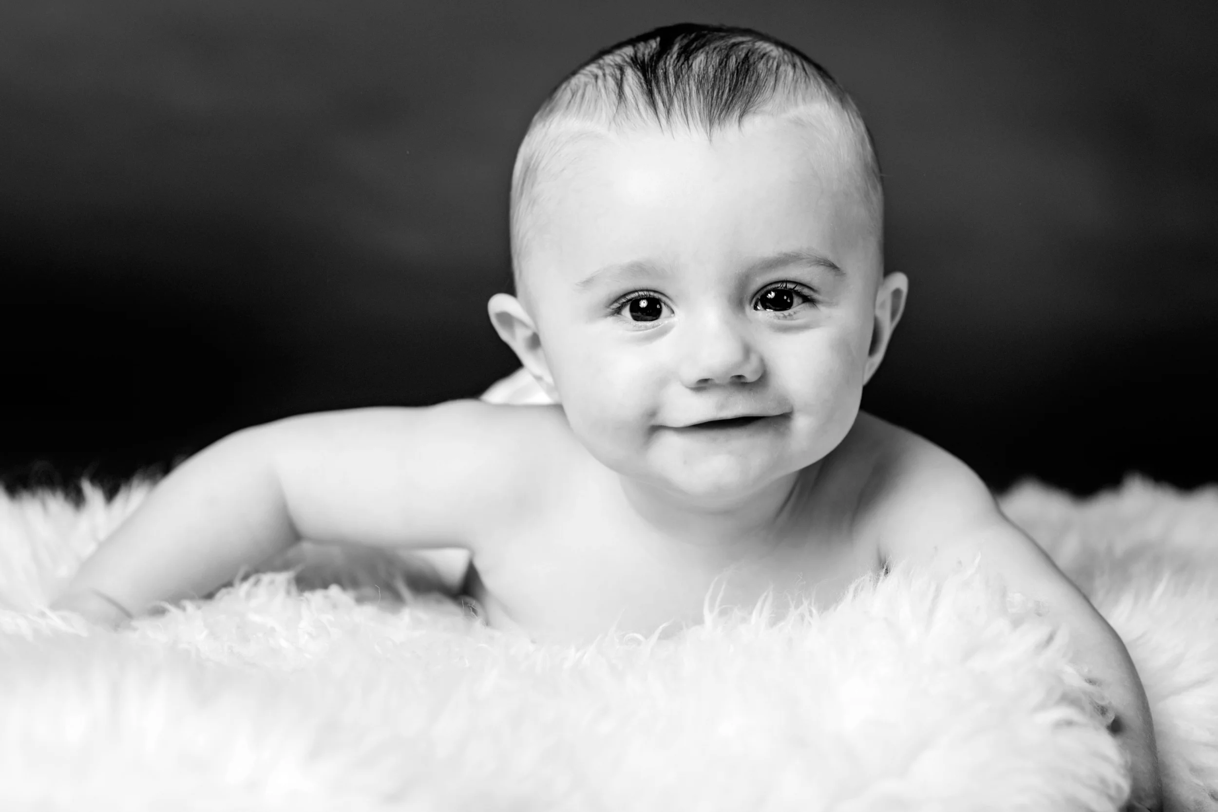 Sort-hvidt babyfoto af glad baby med store øjne, der ligger på maven på et hvidt, blødt tæppe.