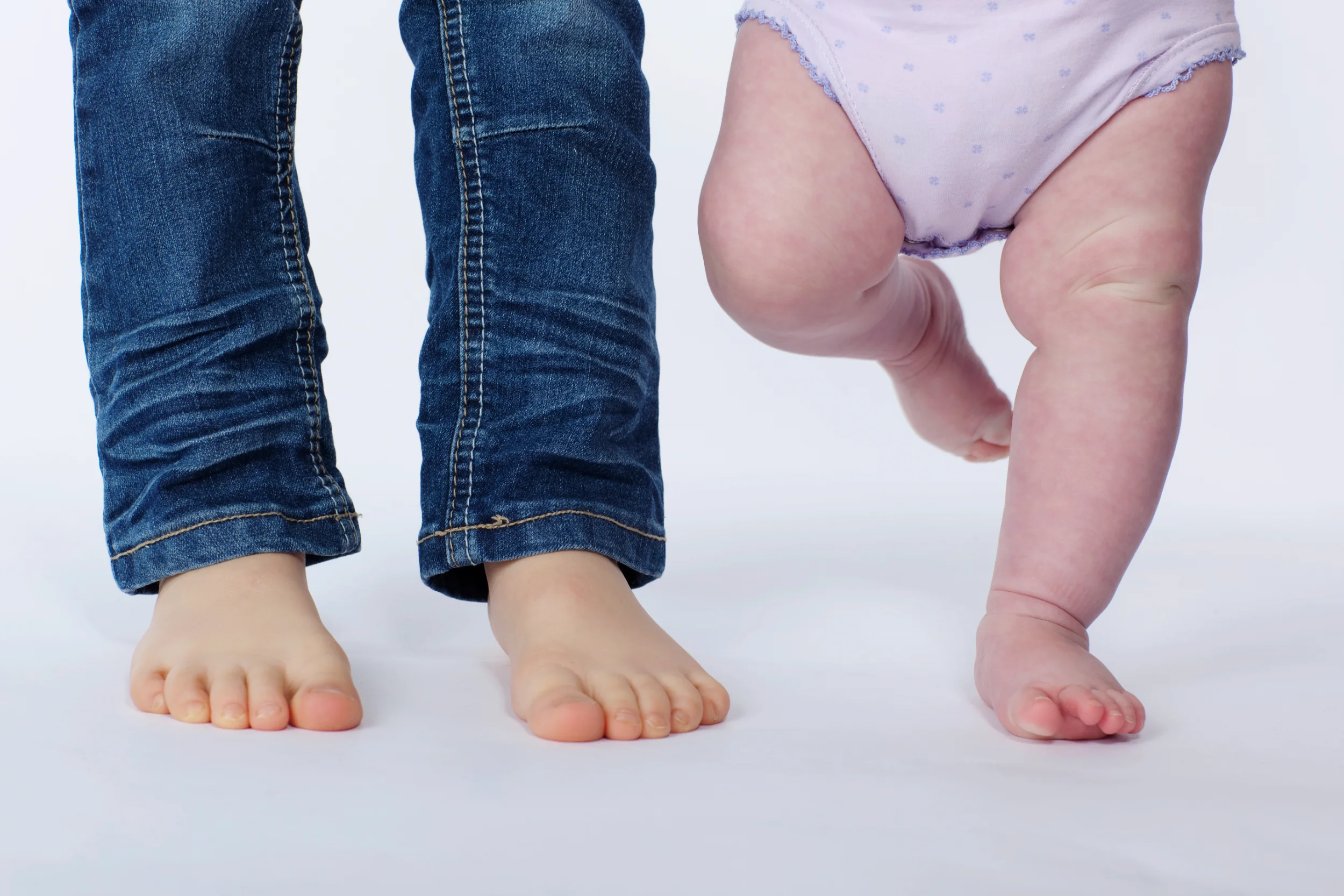 Børnefoto med små bare fødder fra et barn i jeans og en baby i blød lilla ble. Naturligt lys.