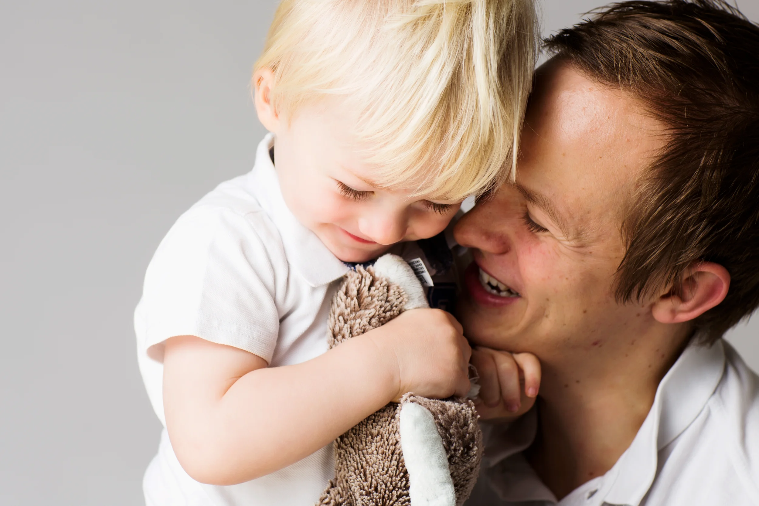 Nært børnefoto af lyshåret barn med tøjdyr, der griner hos sin far mod en lys baggrund. Fanger leg og autentiske smil.