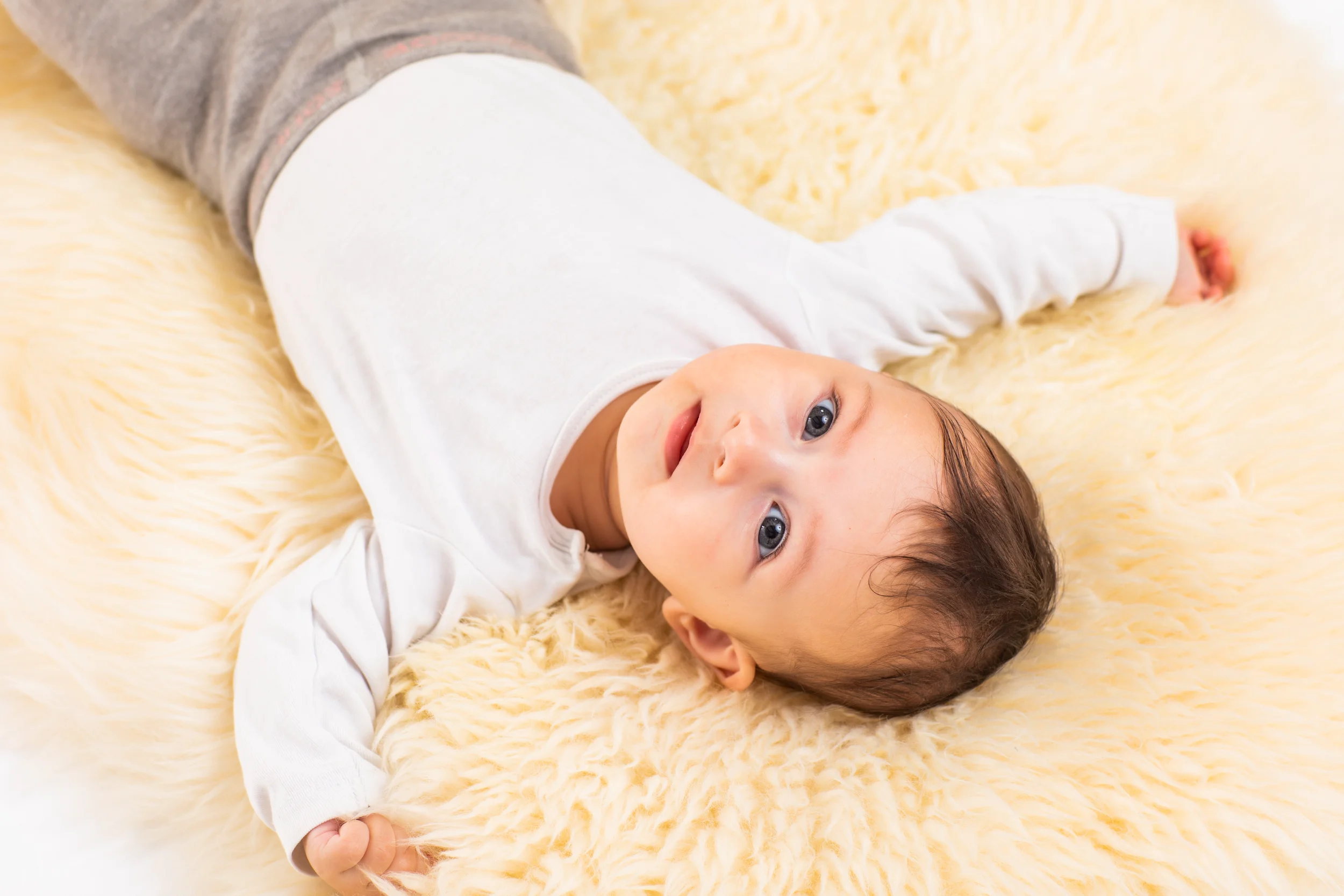 Lille baby med mørkt hår, blåt blik og åben mund liggende på blødt, lyst babylammeskind. Et ægte babyfoto.