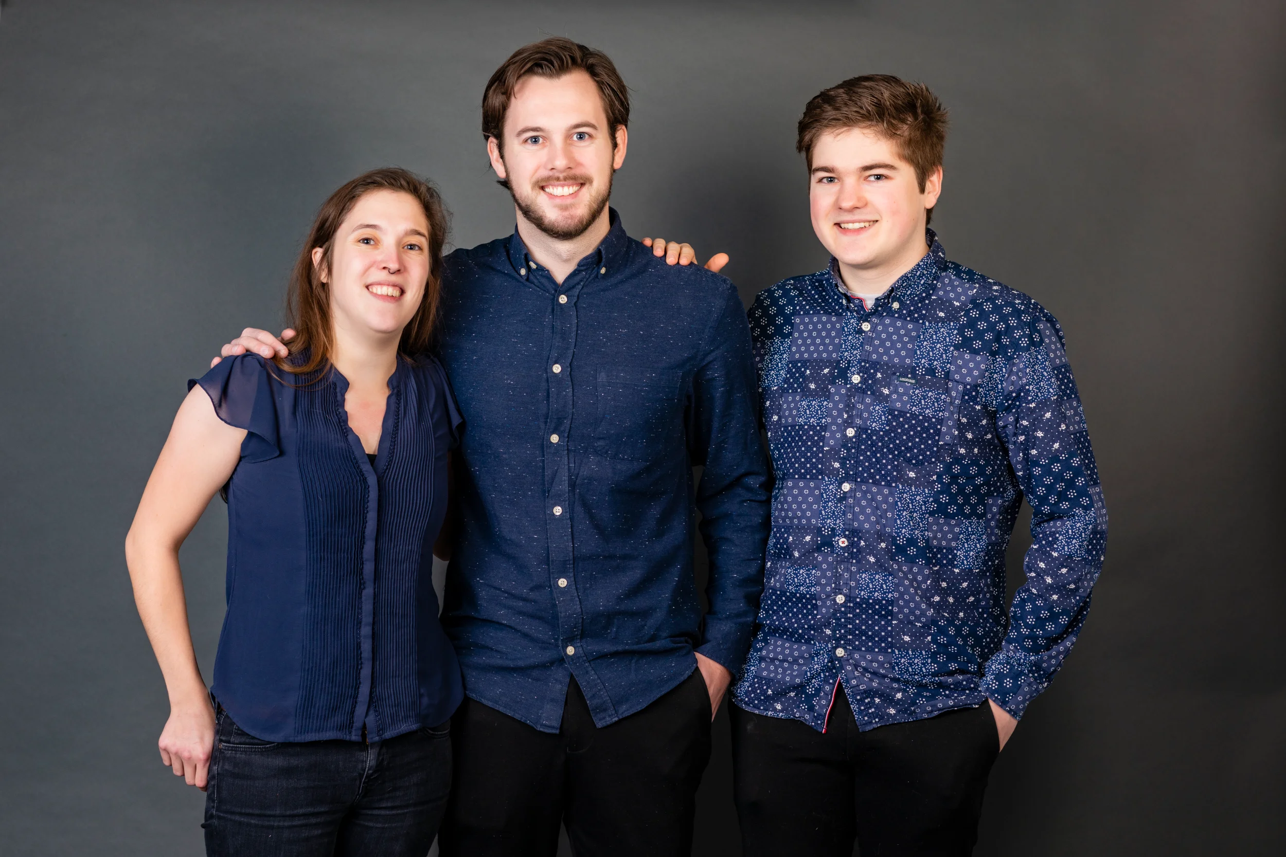 Familiefoto af tre smilende søskende i blåt tøj mod mørk baggrund, som fanger ægte glæde i Virum.