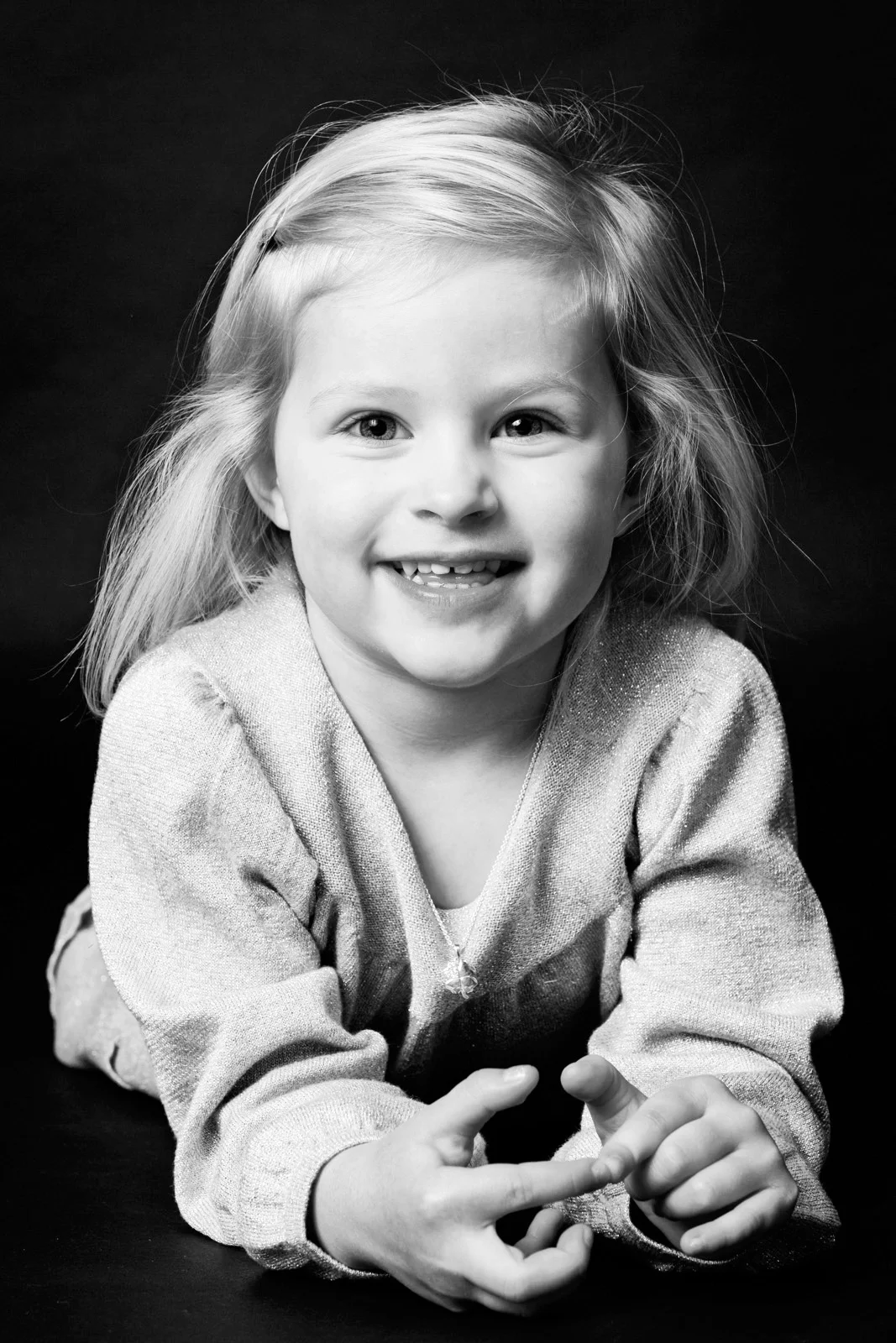 Børneportræt i sort-hvid af smilende pige med lyst hår, der ligger med fremadvendte hænder. Autentisk og glad stemning.