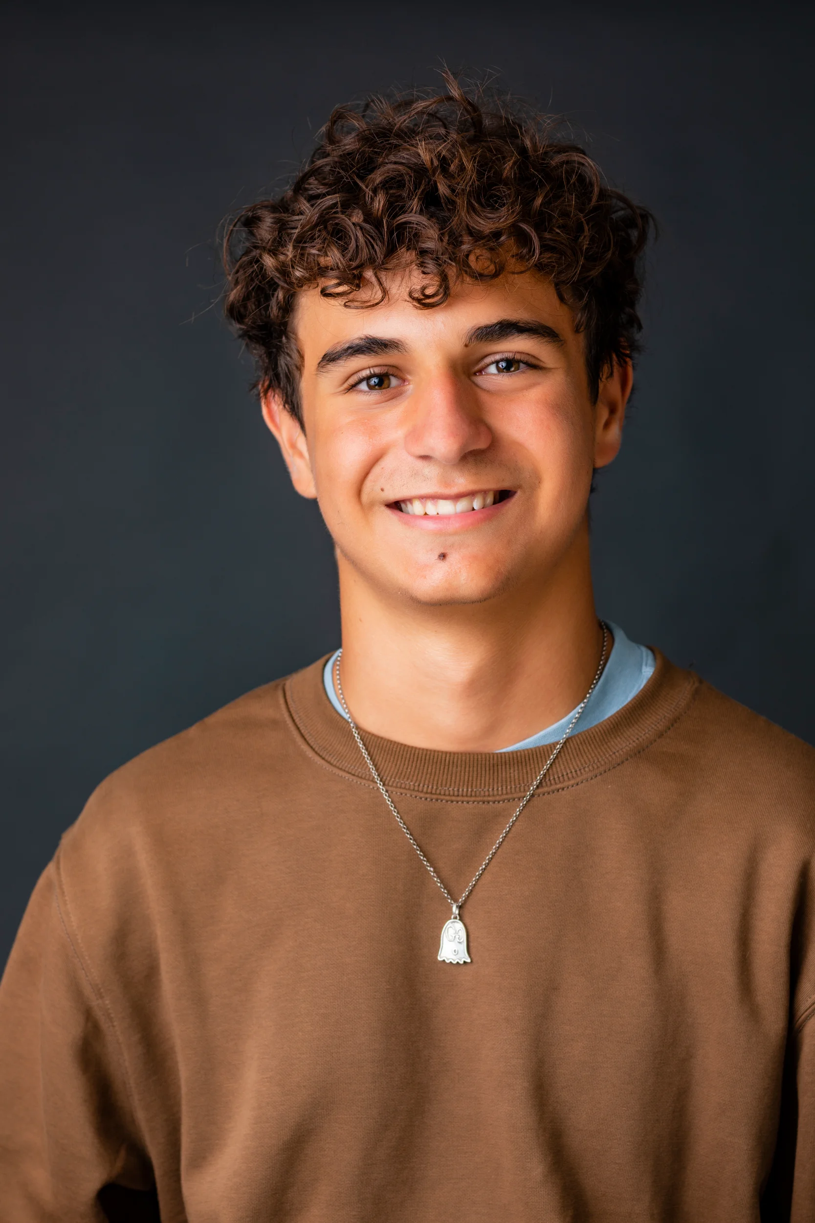 Sort-hvidt portrætfoto af smilende ung mand med krøllet hår og halskæde mod mørk baggrund. Venligt udtryk.