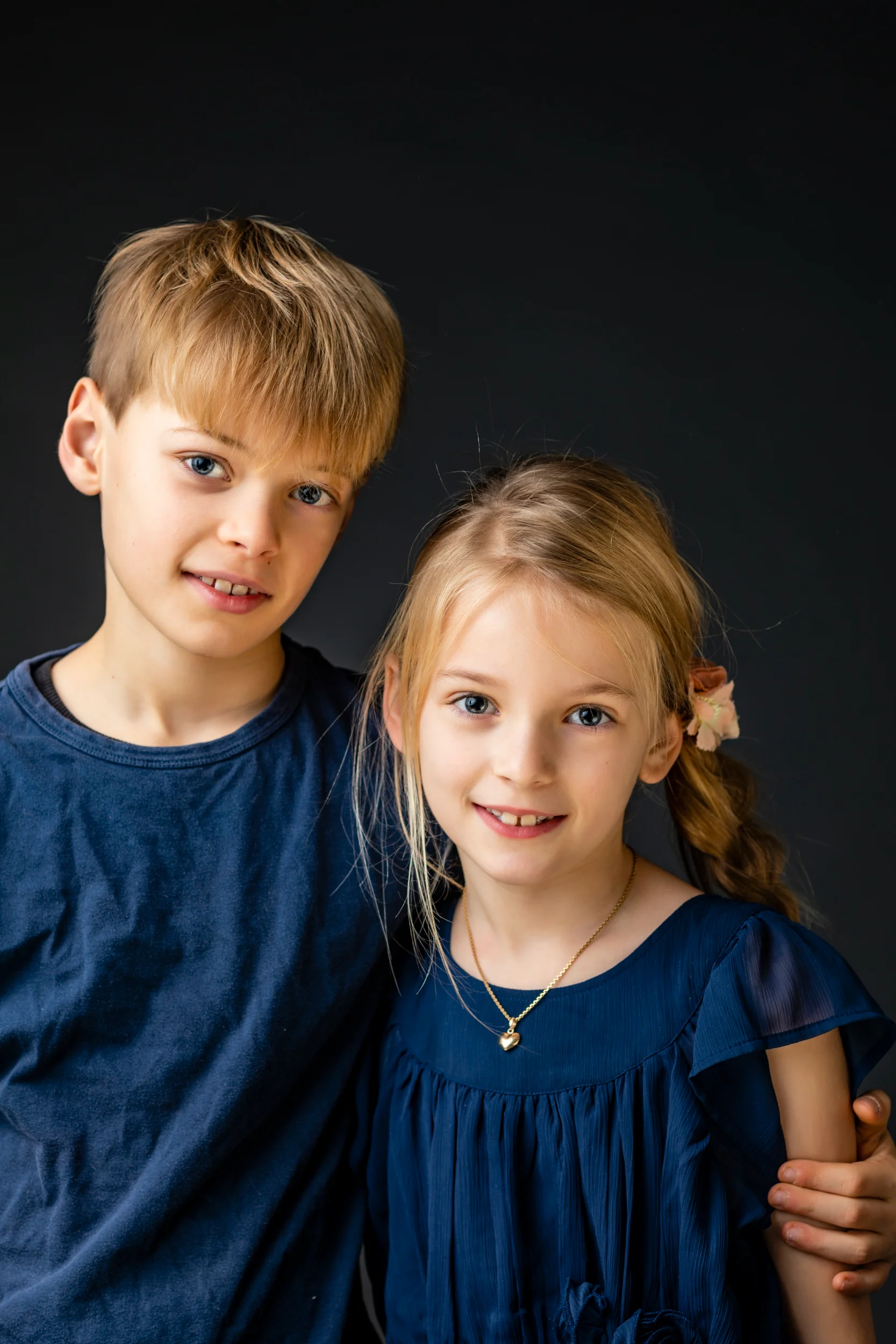 Børnefotografering: Søskende i blåt tøj smiler mod kameraet, mod mørk baggrund i studie, autentisk portrætfoto af børn.