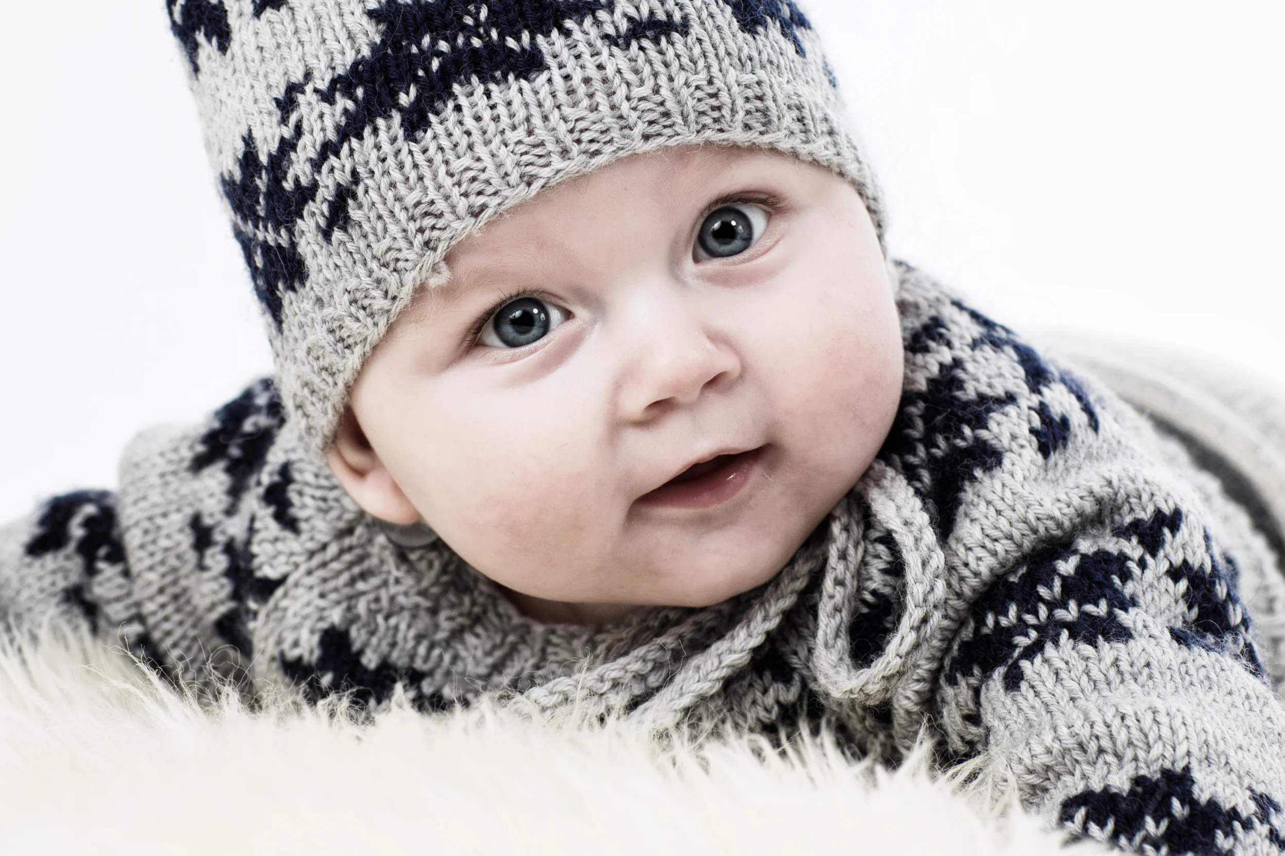 Sødt babyportræt i Virum med blå øjne i strikhue og trøje, der smiler blidt mod beskueren. Lyst, venligt lys.