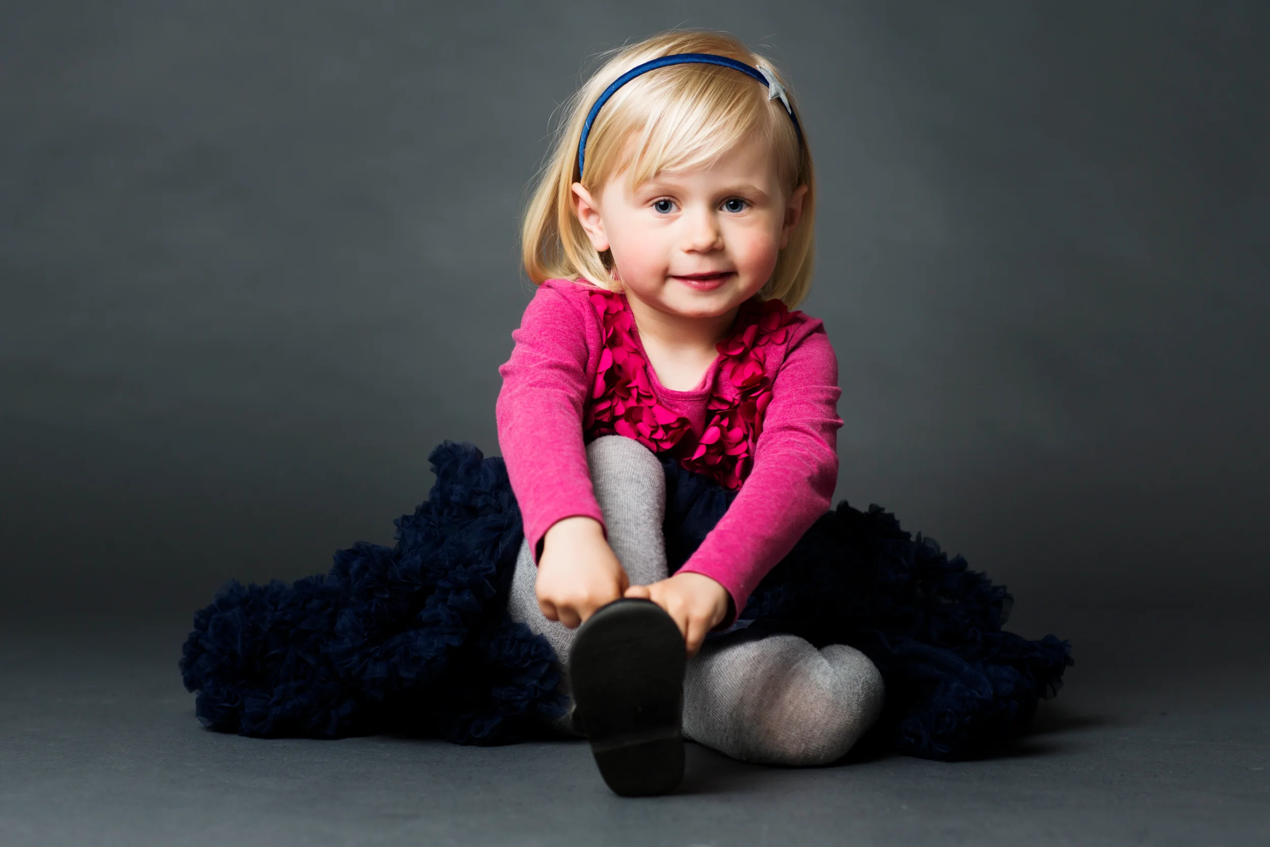 Børnefotografering af blond smilende pige i lyserød bluse og mørkeblåt skørt, der sidder på gulvet.