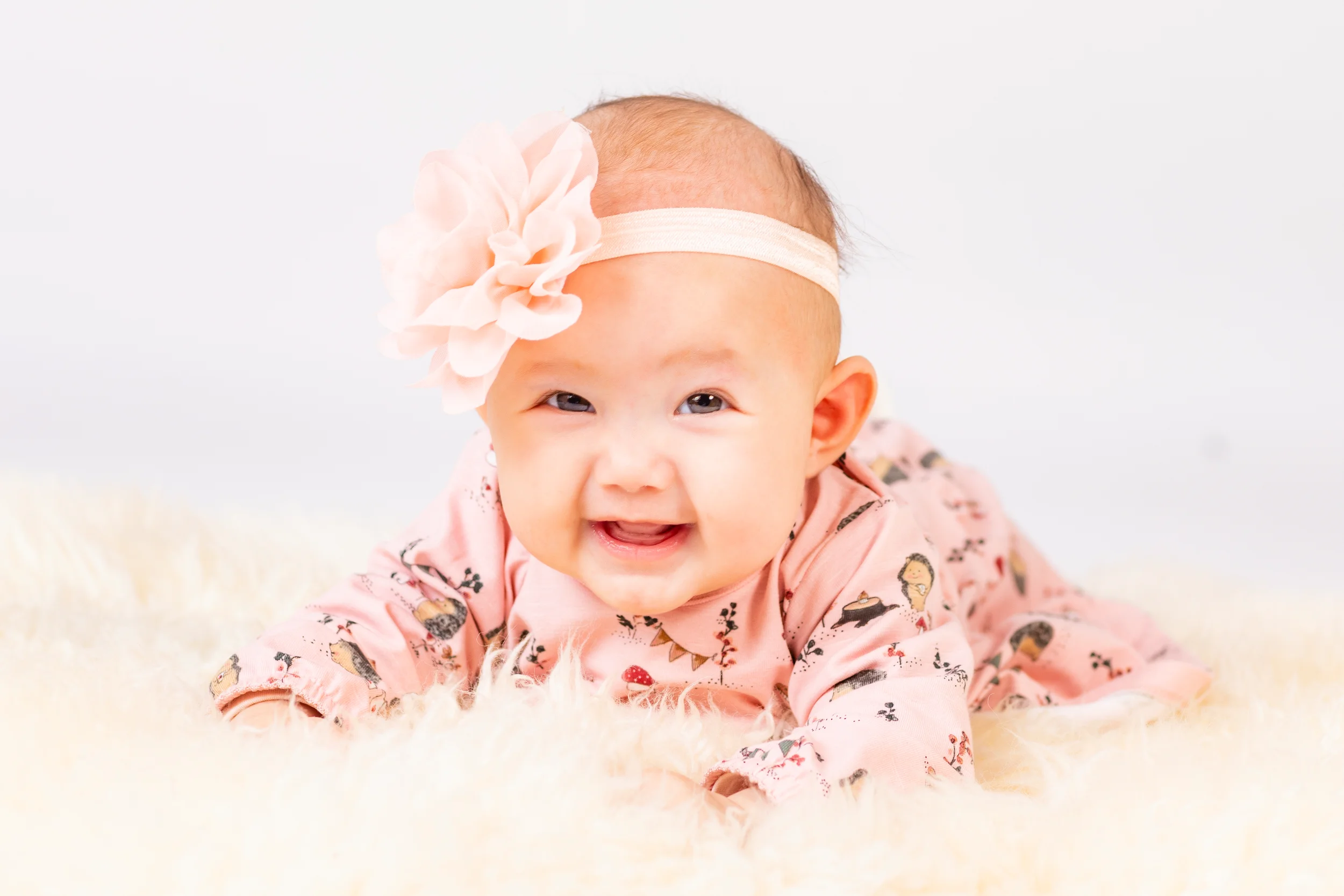 Baby smiler glad iført lyserød heldragt med dyremotiver og lyserødt hårbånd med blomst, liggende på blødt, hvidt tæppe. Ba...