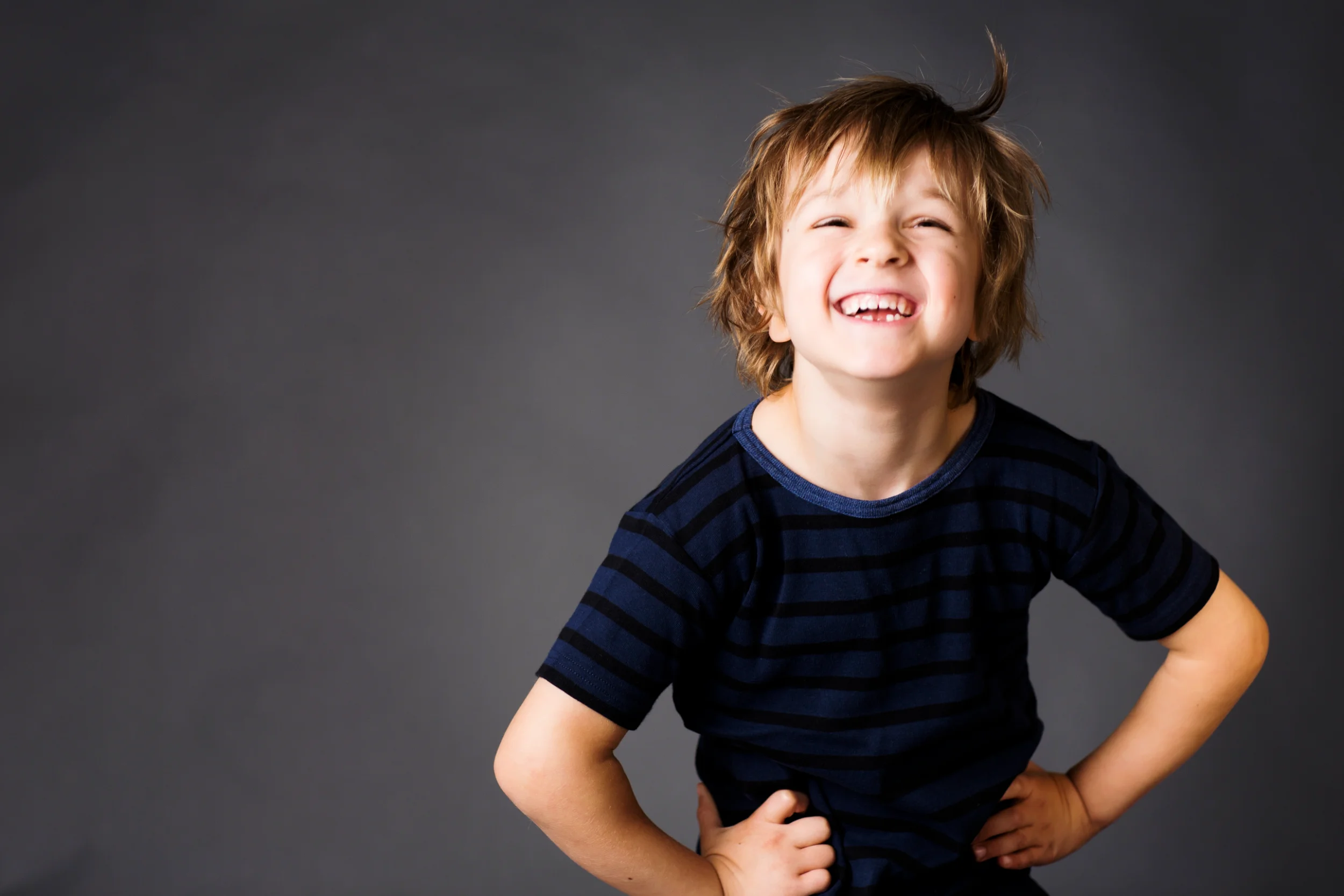 Børnefoto af en glad, legende dreng med stort smil, rodet hår og hænderne i siden på en ensfarvet grå baggrund.