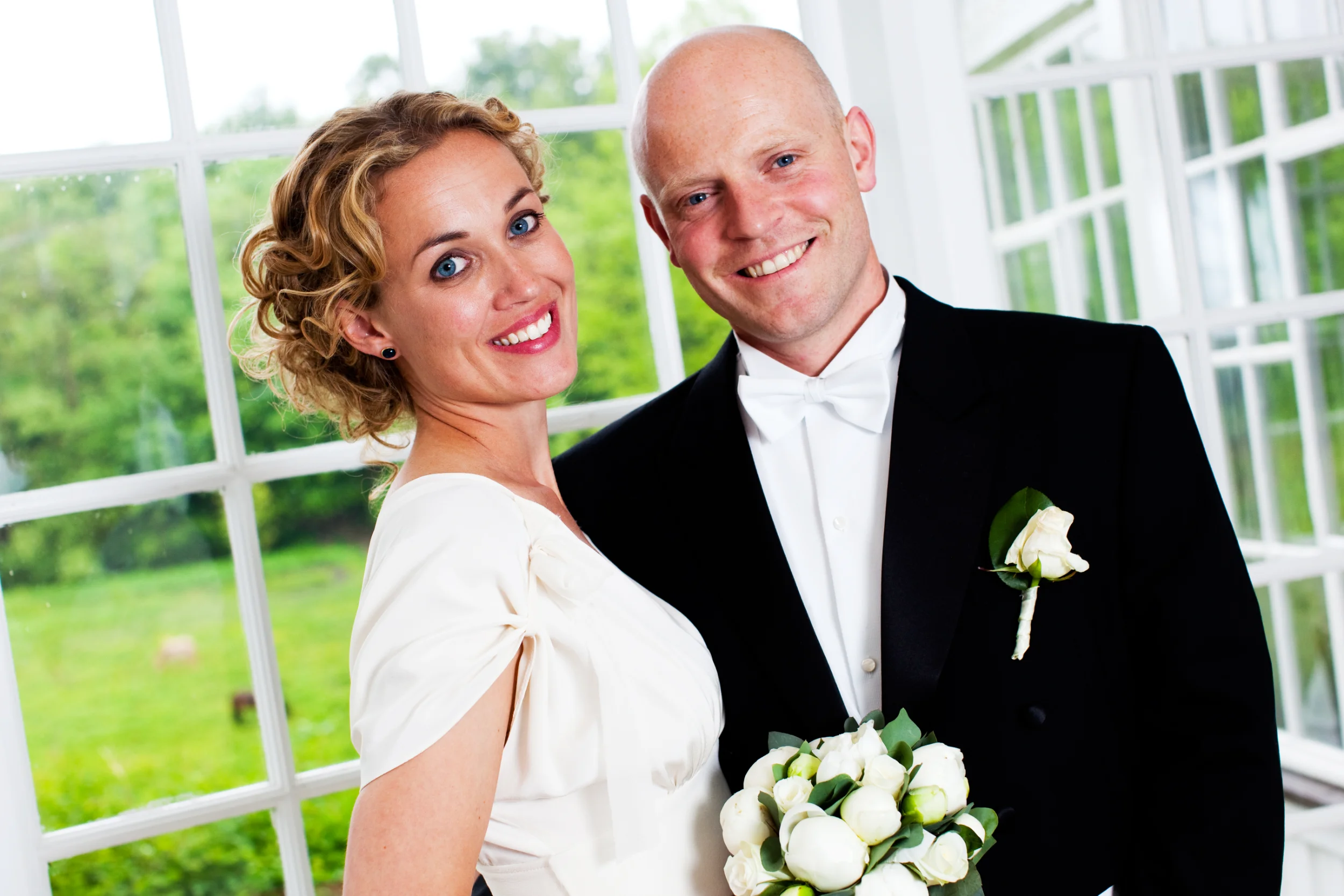 Bryllupsfoto af smilende par med brudebuket, stående elegant foran et vindue med grønne træer i baggrunden.