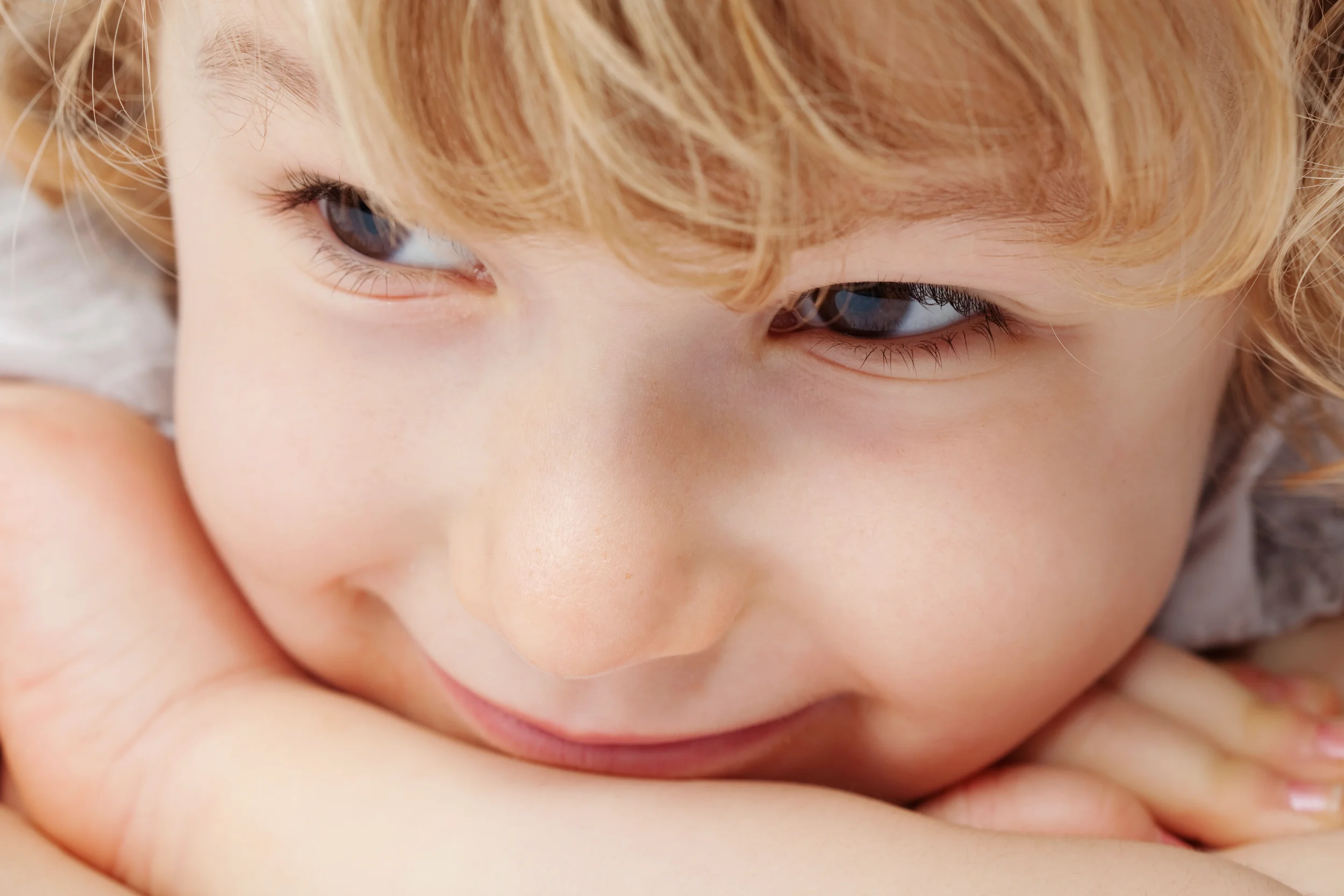 Nærbillede af et lyst, smilende barn med nysgerrige øjne og lyse krøller, der fotograferes i et blødt lys. Fokus på barns ...