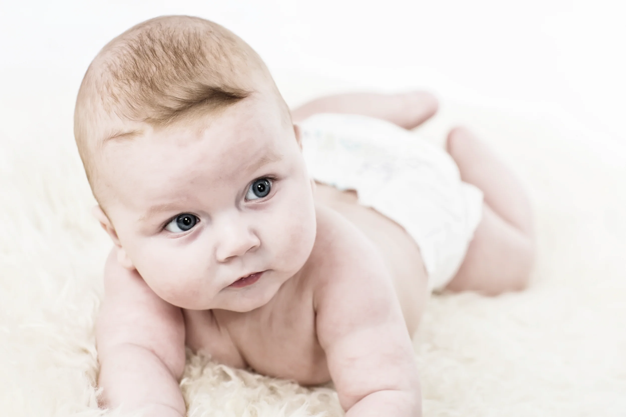 Babyfotografering: Nysgerrigt babyportræt med blå øjne, liggende på lyst tæppe og kigger op mod beskueren i blødt lys.