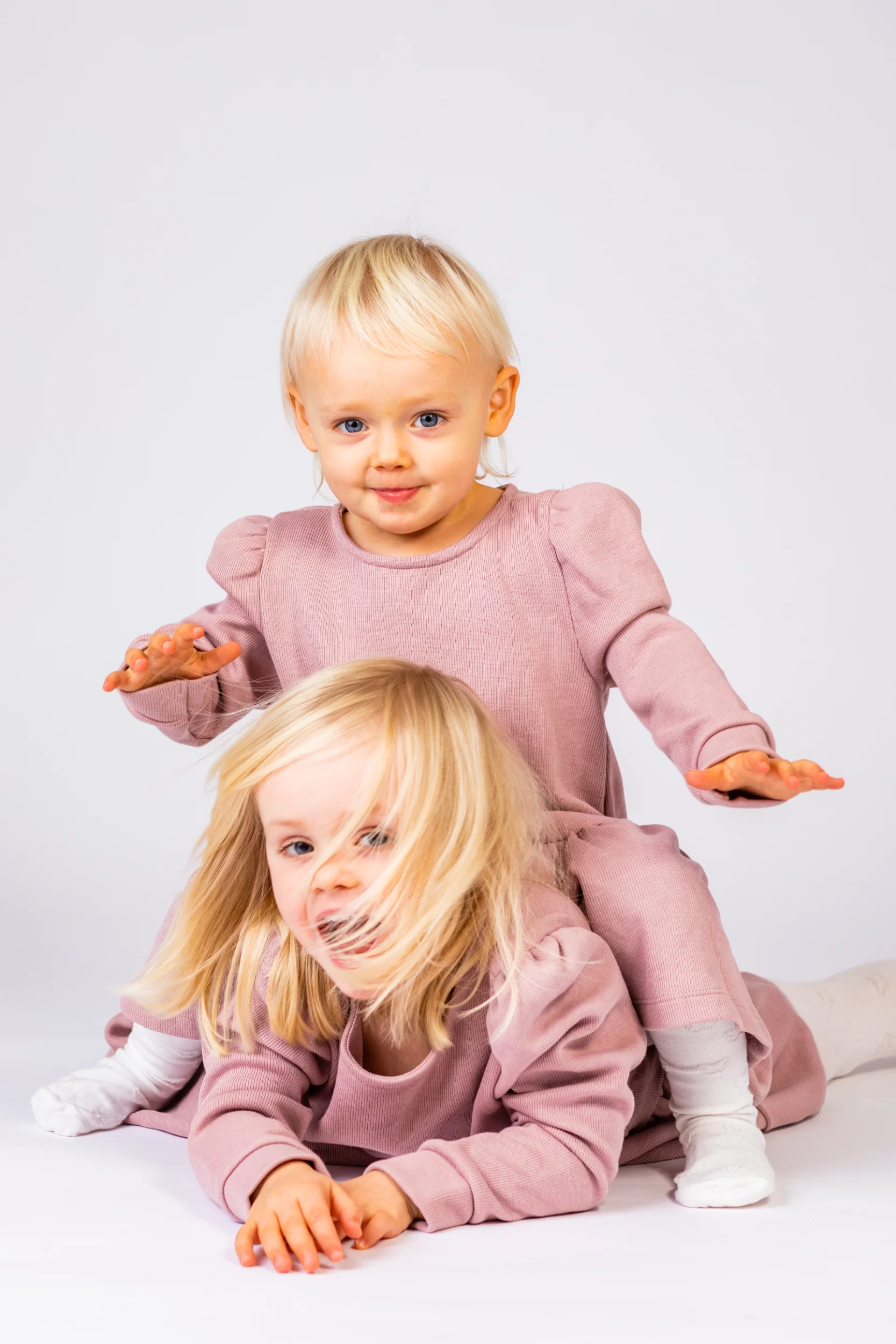 Legesyge børnebilleder: To smilende små piger med lyst hår i lyserøde sæt, den ene kravler på den anden.