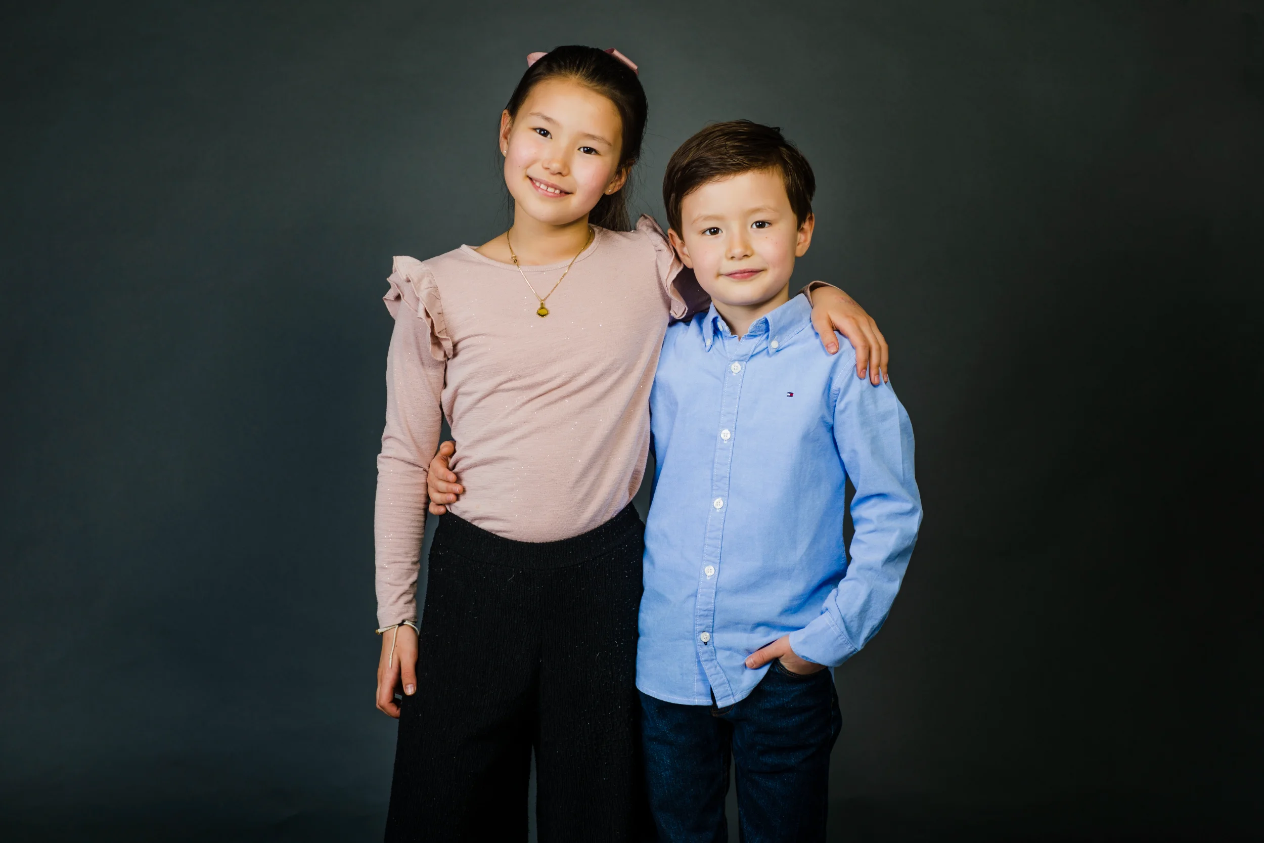 Børn: Søskendeportræt af to smilende børn, en pige og en dreng, poserer tæt sammen foran en mørkegrå baggrund.