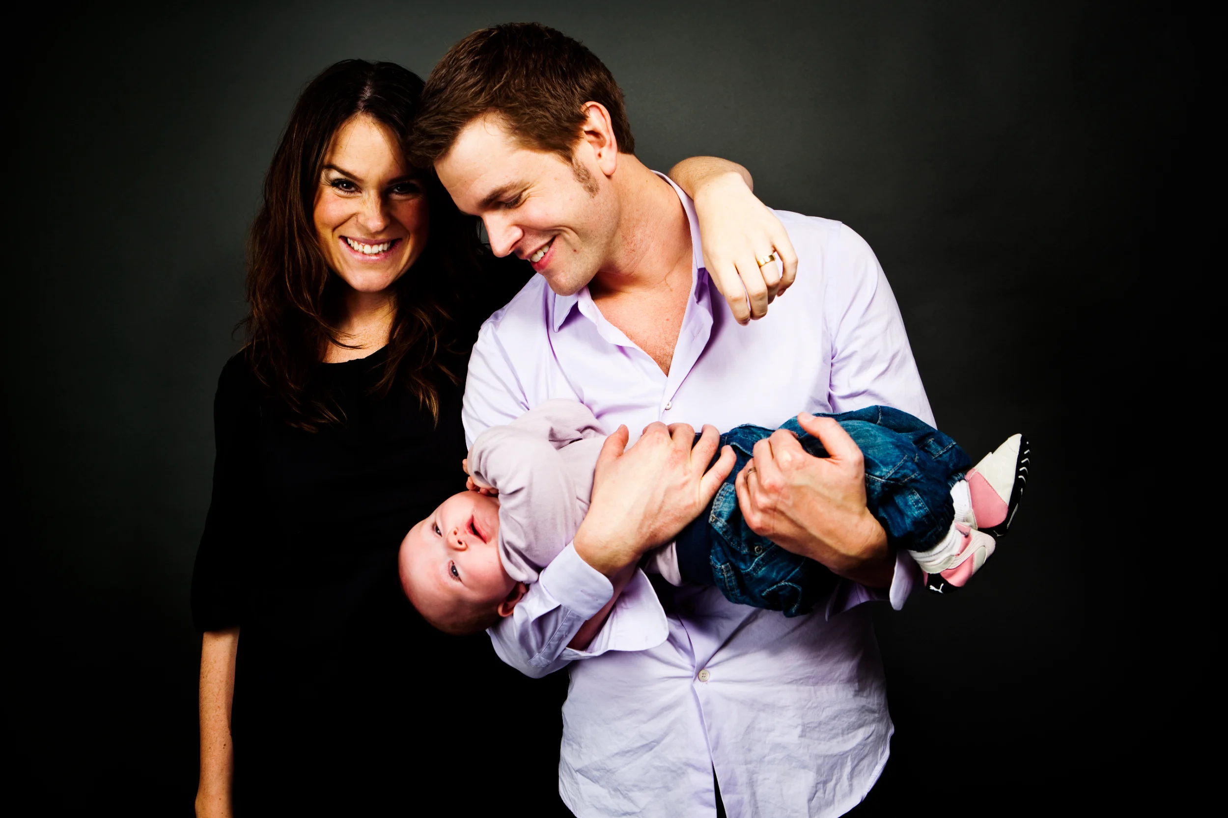 Familiefoto: Forældre smiler varmt, mens far holder baby glædeligt på hovedet mod en mørk baggrund.