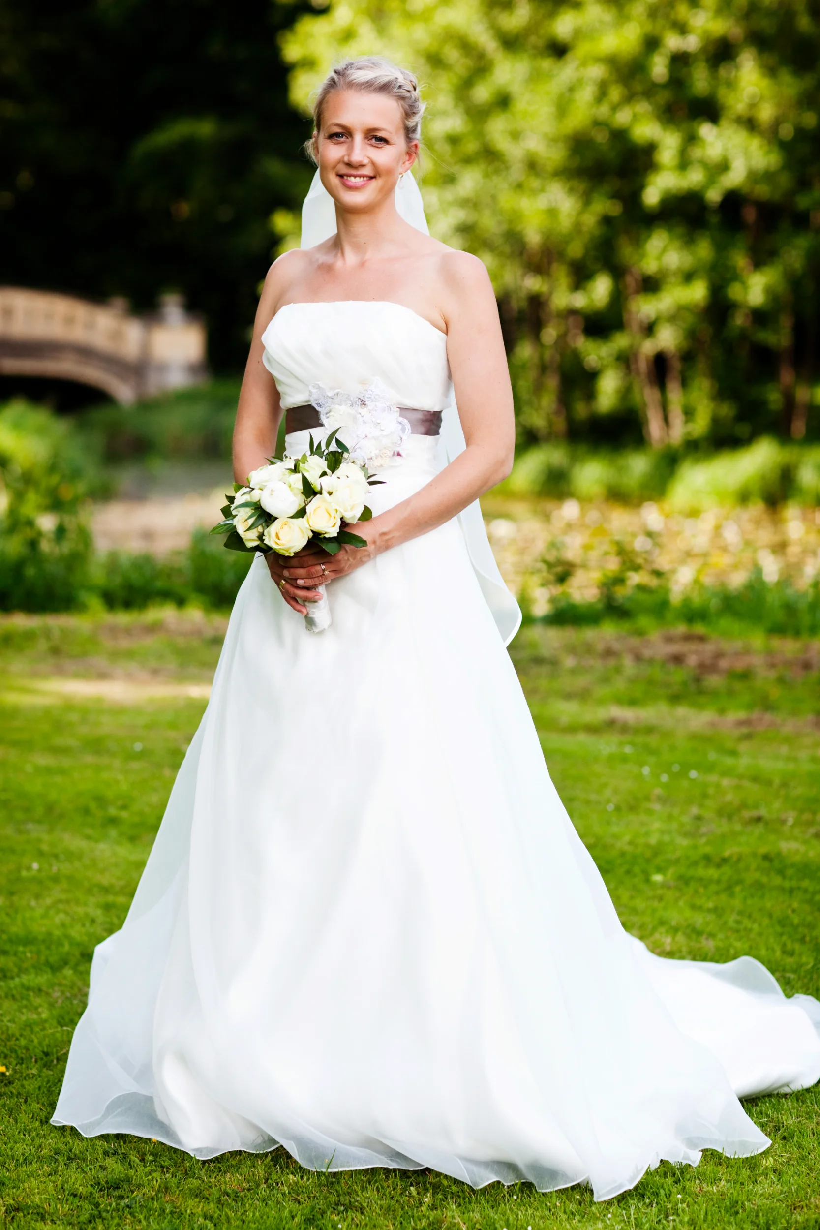 Bryllupsfotografering af glad brud i hvid kjole med brudebuket, stående på græs i Aldershvile slotspark.