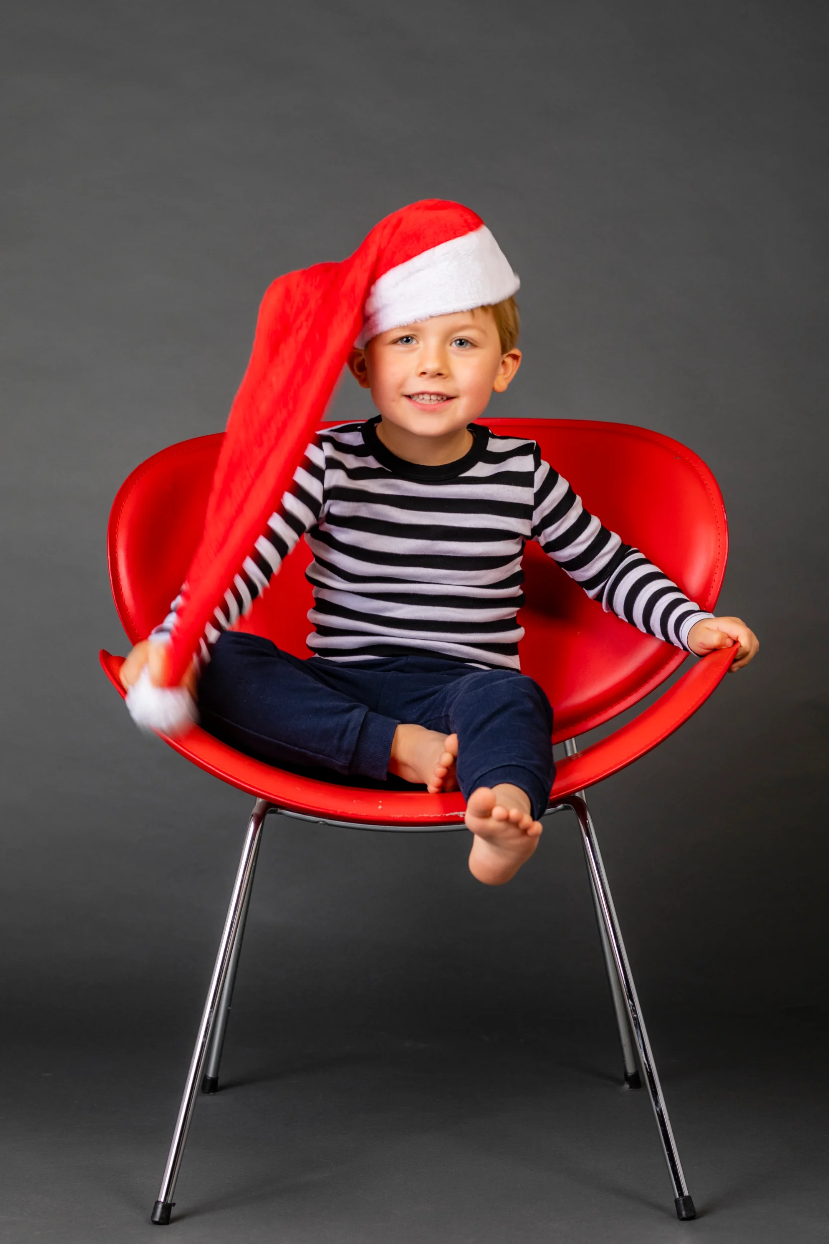 Glad dreng med nissehue i personligt børnefotografi. Drengen sidder på rød stol, lyserøde kinder og smiler bredt.
