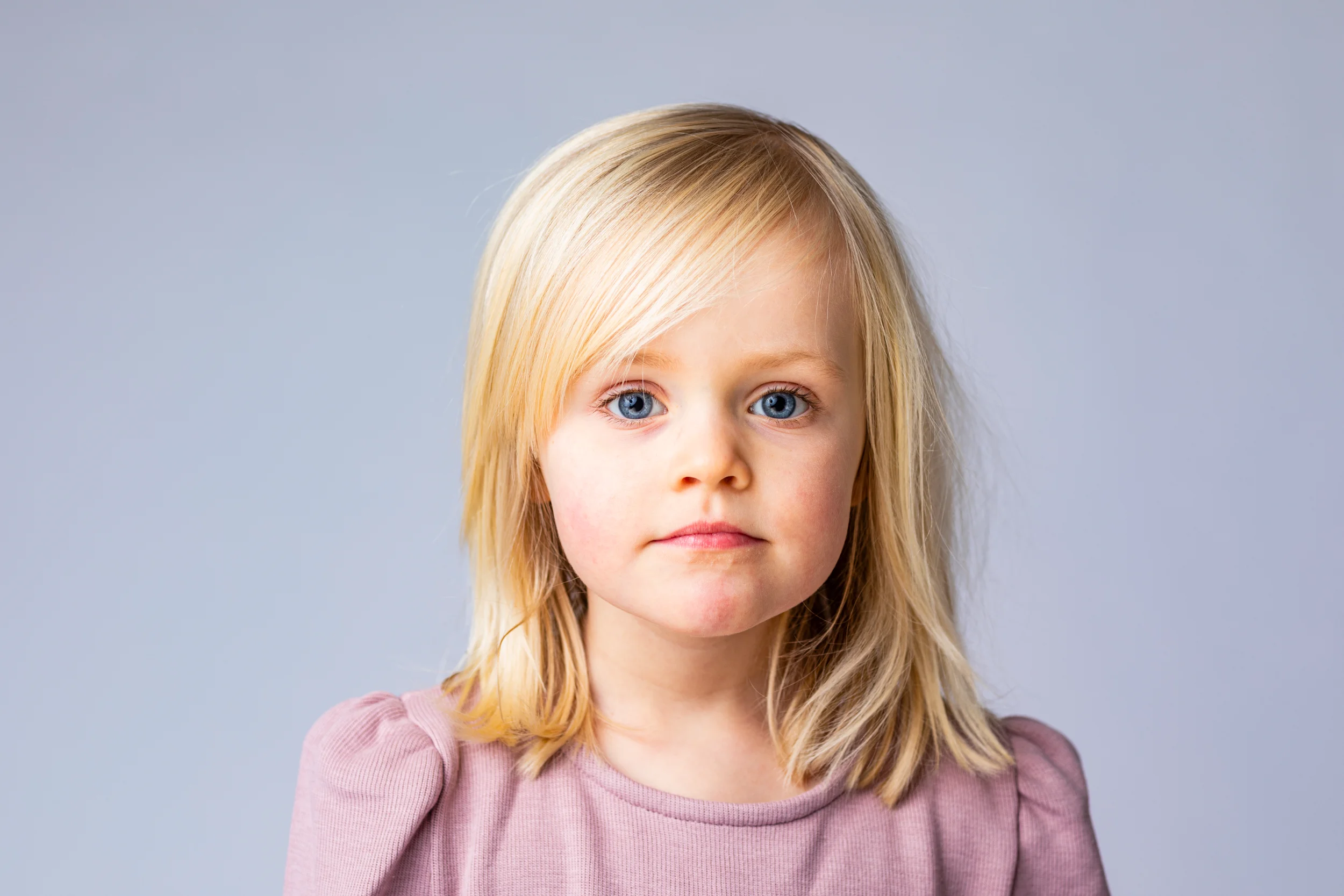 Børnefotografering af pige med blondt hår og intense blå øjne, neutral baggrund.