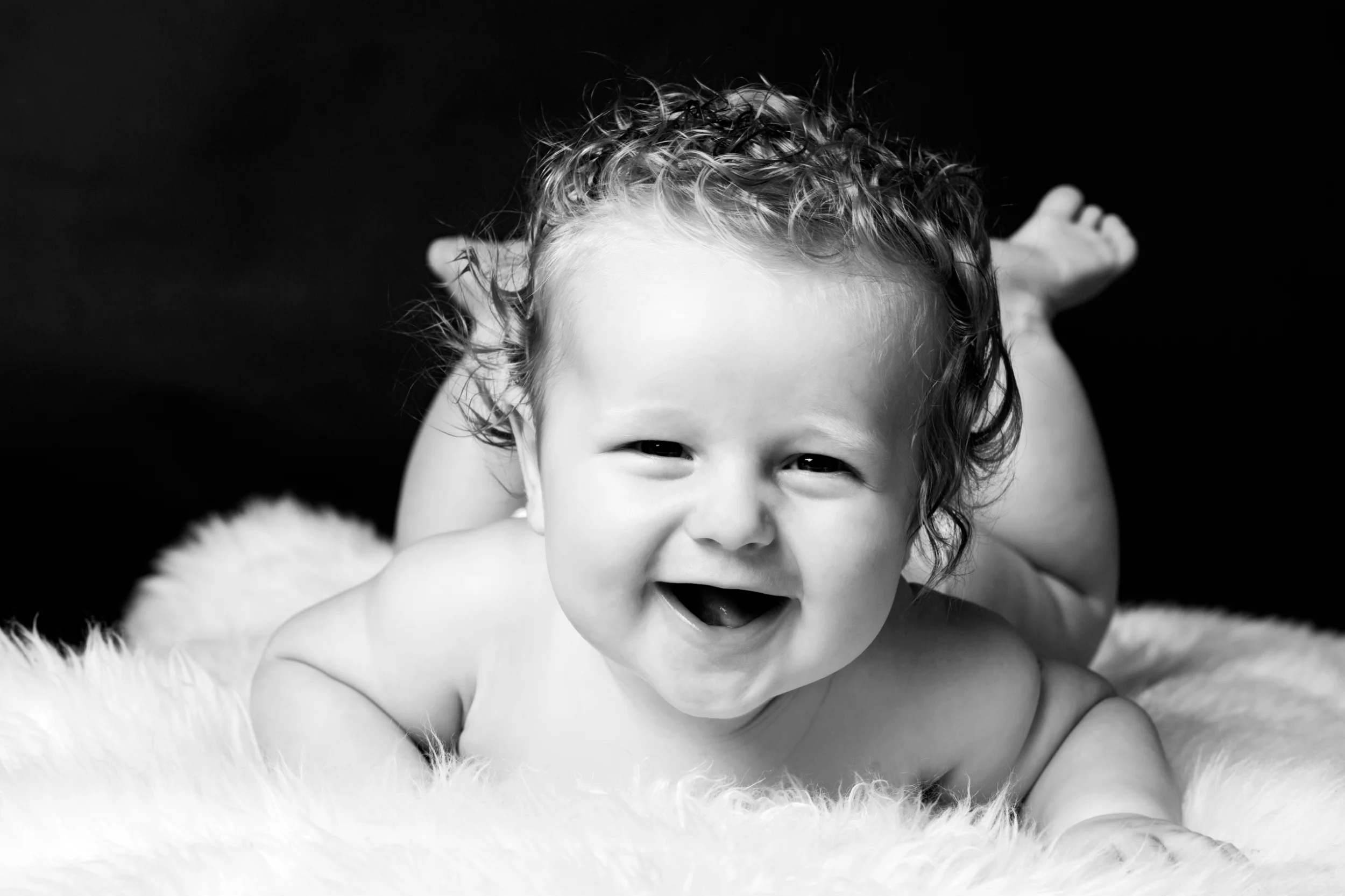 Sort/hvidt babyfotografi af en glad baby der smiler stort, liggende på maven på et hvidt tæppe.