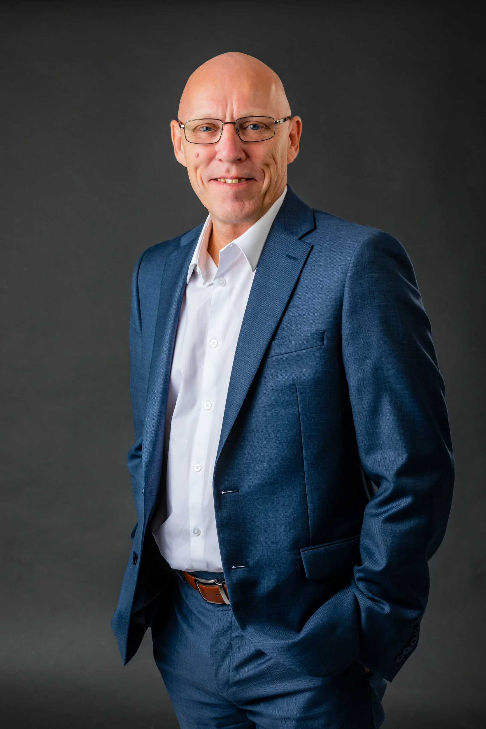 Glad mand i blåt jakkesæt og hvid skjorte med hænder i lommerne. Professionelt studieportræt i dagslys.